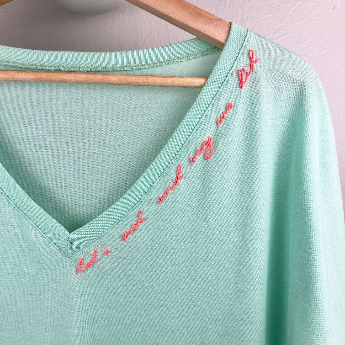 close up view of a neckline of a mint green v-neck tee with hand embroidery along the neckline that says "let's not and say we did" in neon pink thread