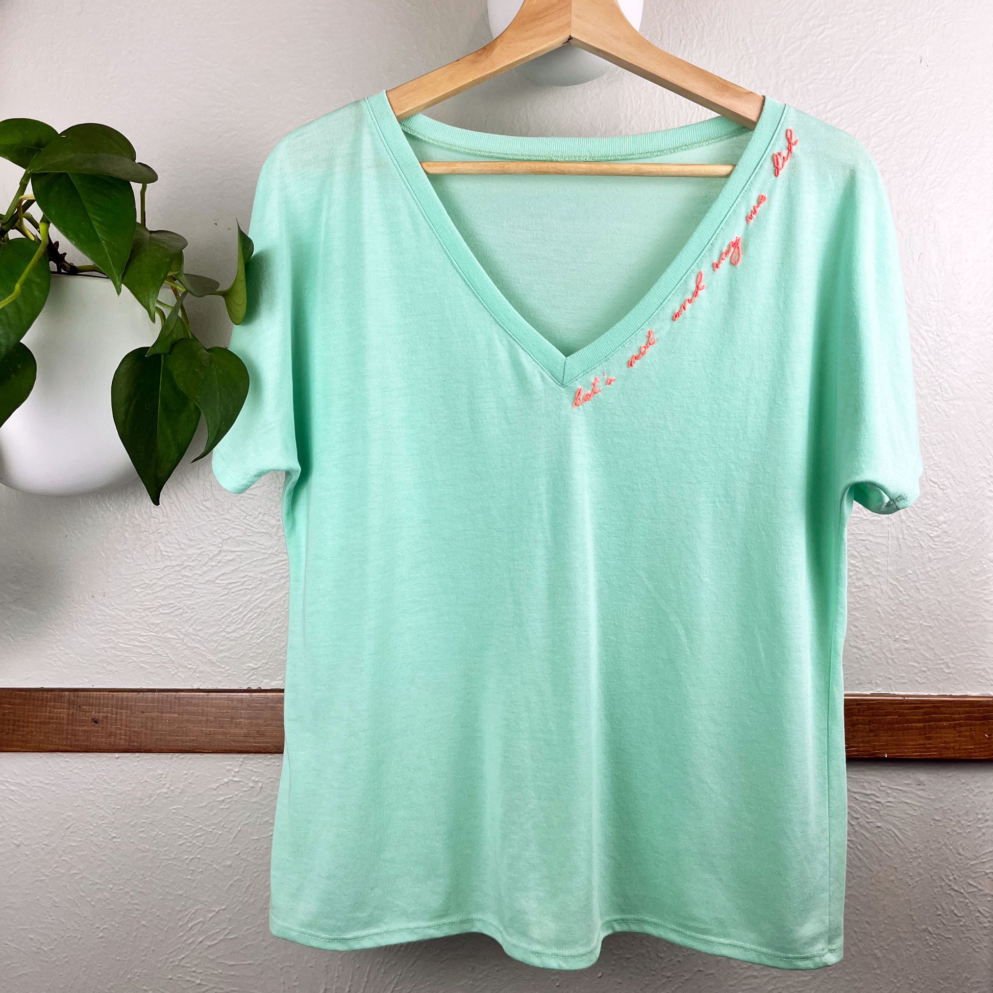 mint green v-neck tee with hand embroidery along the neckline that says "let's not and say we did" in neon pink thread, hanging on a wall next to a pothos plant