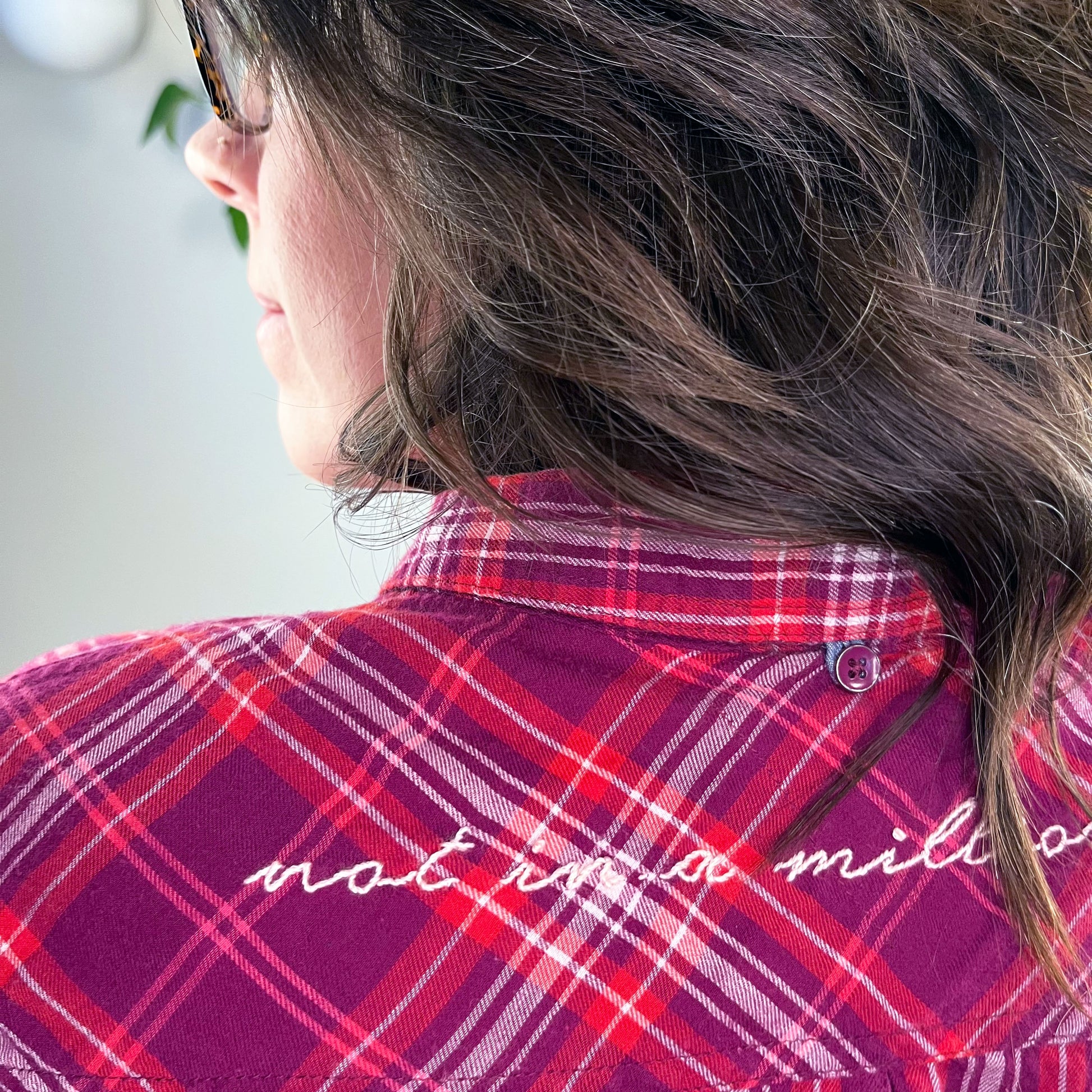 close up view of the back shoulders of a woman wearing a berry colored plaid flannel shirt with scripty words not in a million hand embroidered on the back yoke