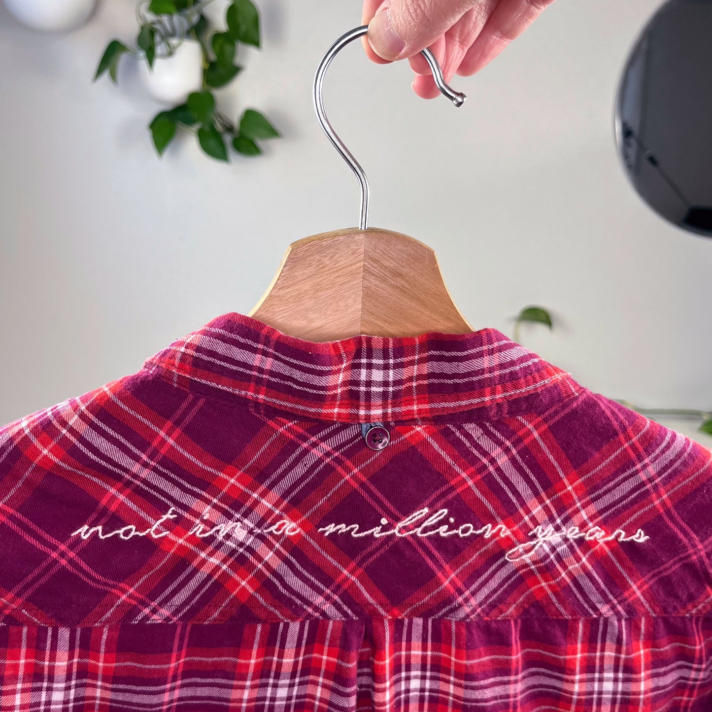 back close up view of a berry colored plaid flannel shirt on a hanger with scripty words not in a million years hand embroidered on the back yoke