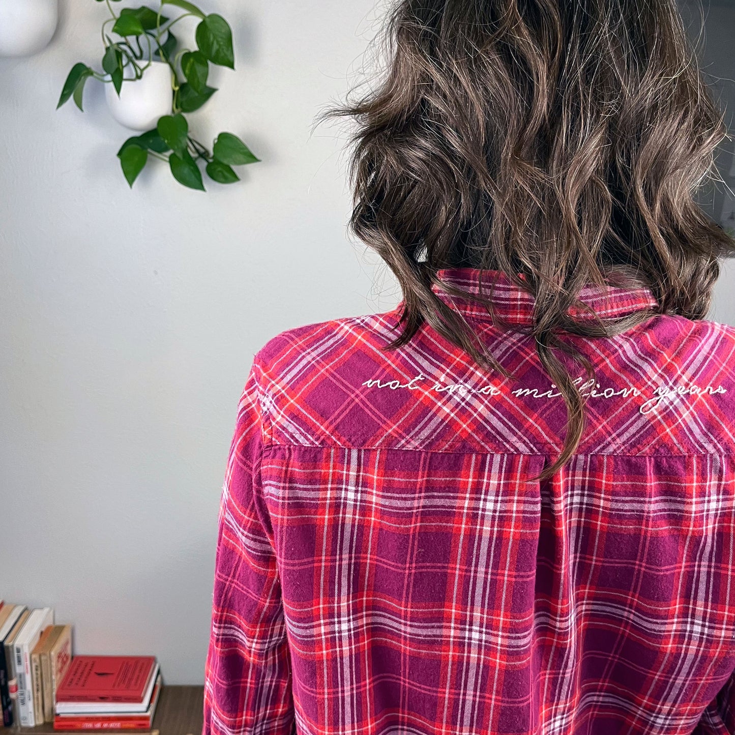 back view from the waist up of a woman wearing a berry colored plaid flannel shirt with scripty words not in a million years hand embroidered on the back yoke