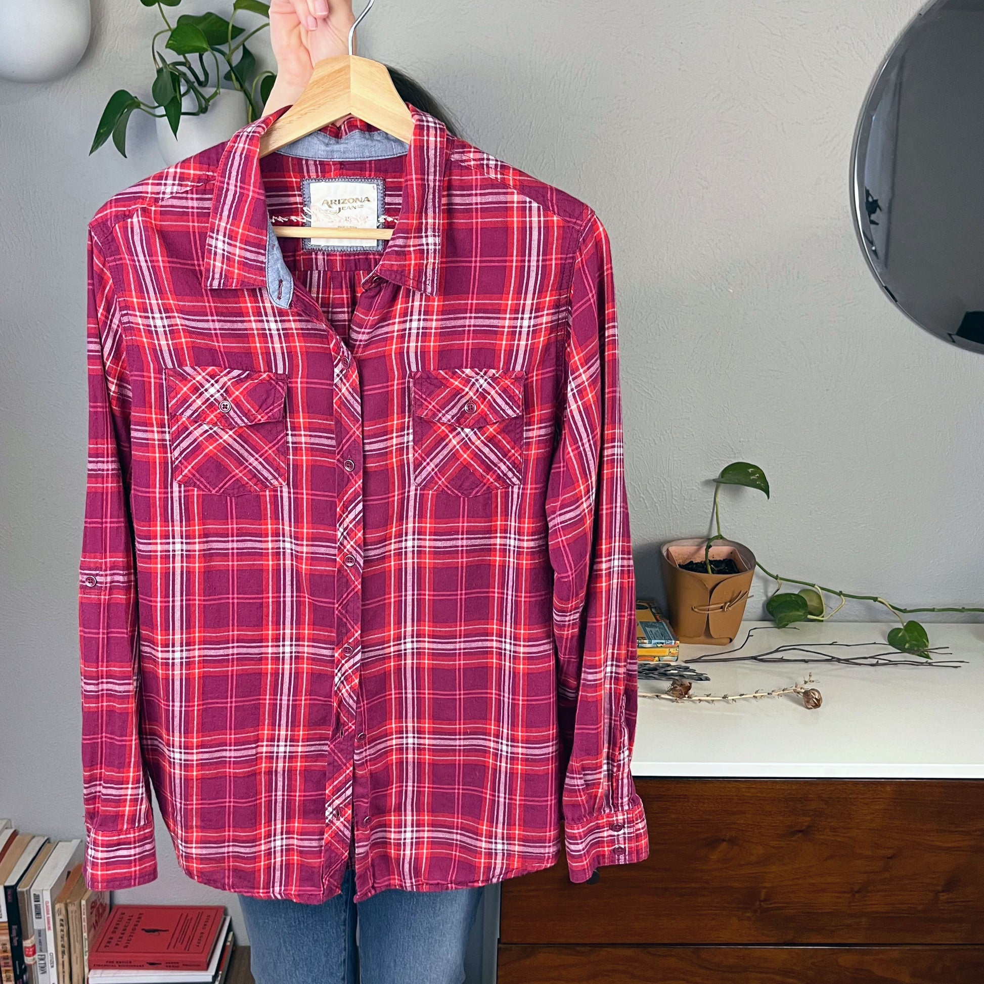 front view of a berry colored plaid flannel shirt on a hanger being held up by.a person you can't see in front of a few pothos plants