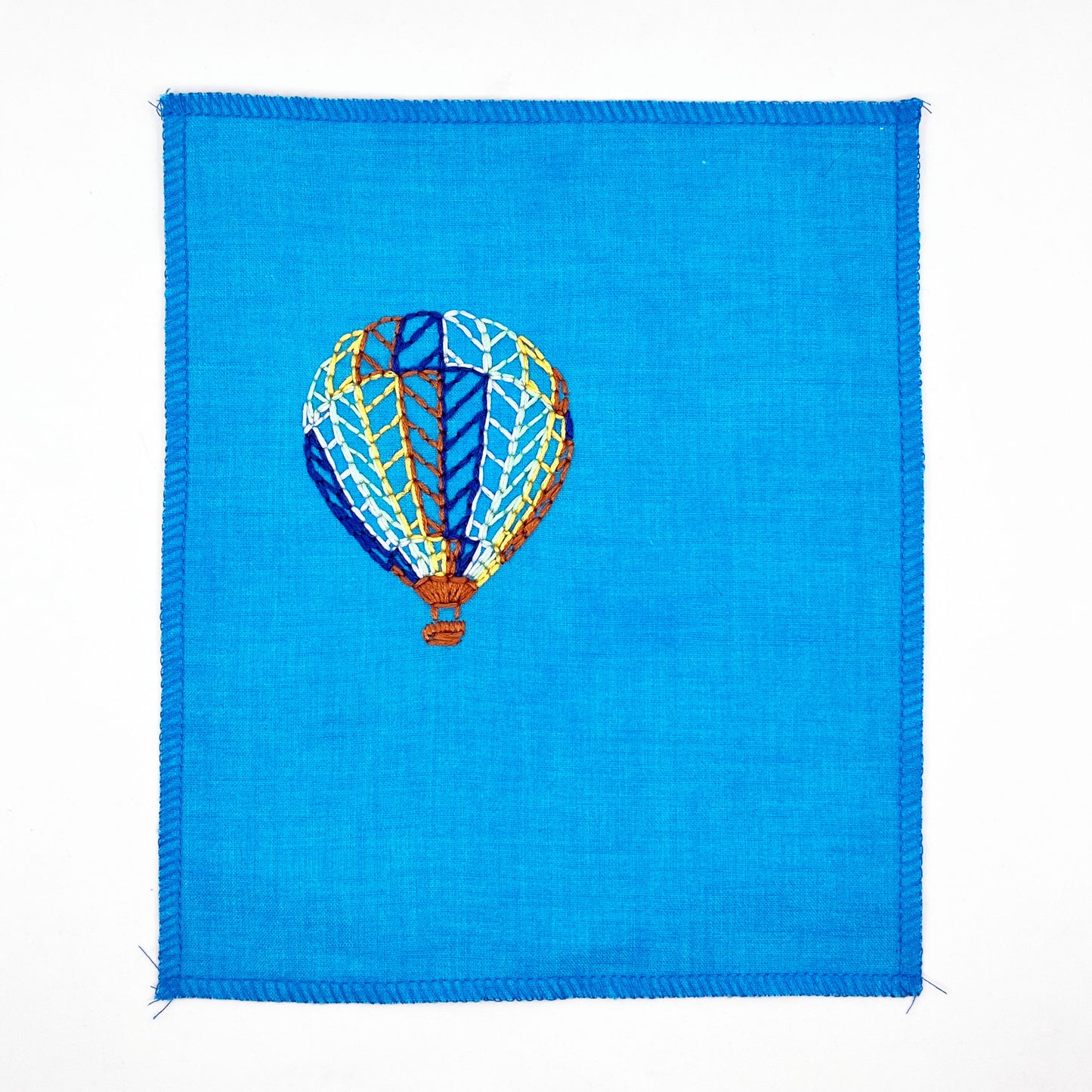 a bright blue fabric wall hanging with a hot air balloon stitched on it off center, in dark blue, light blue, light green, yellow and rust colored thread, on a white background