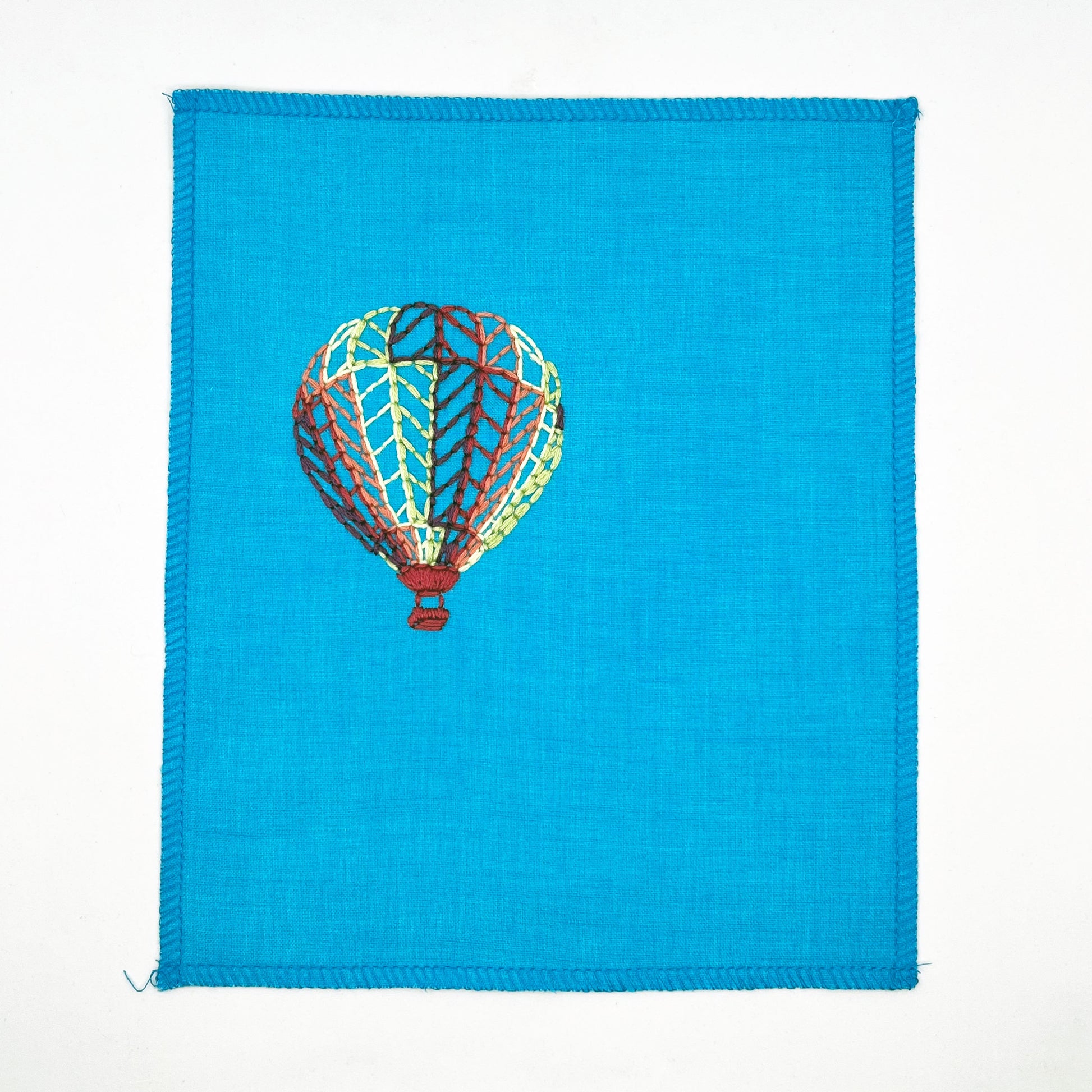 a bright blue fabric wall hanging with a hot air balloon stitched on it off center, in plum, magenta, coral, lime green and yellow colored thread