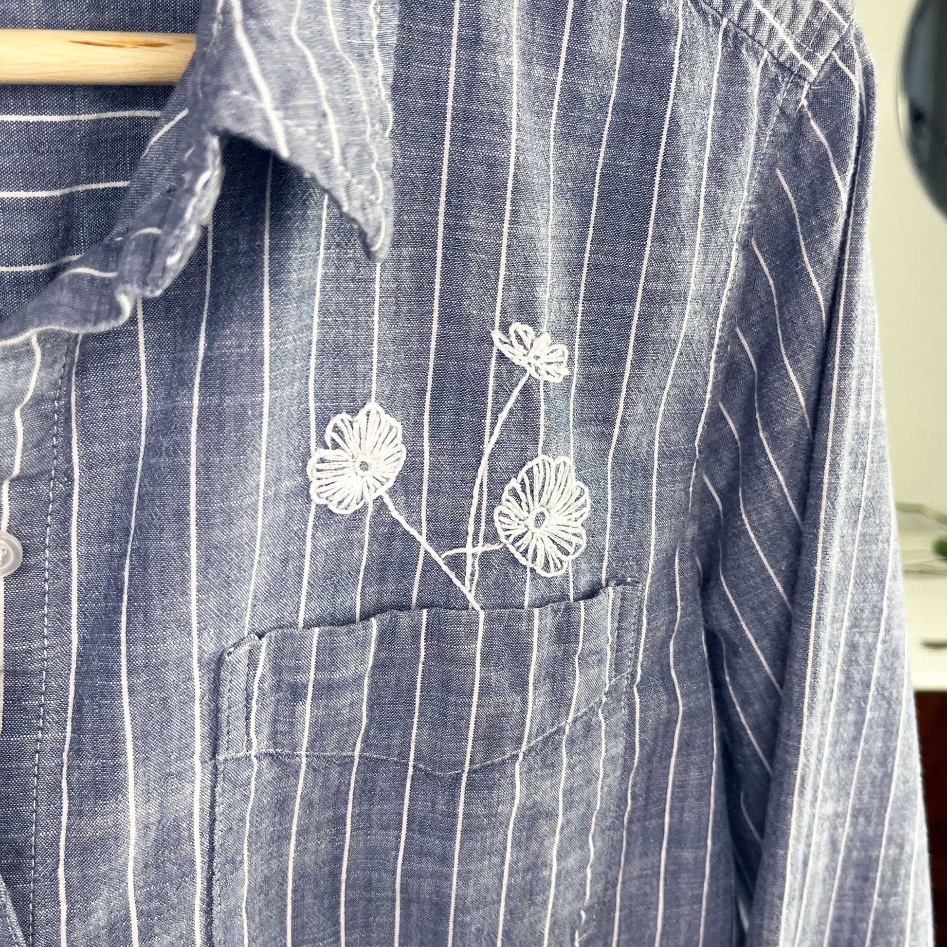 close up view of a striped chambray shirt with white hand embroidered poppies coming out of the chest pocket being held up on a hanger