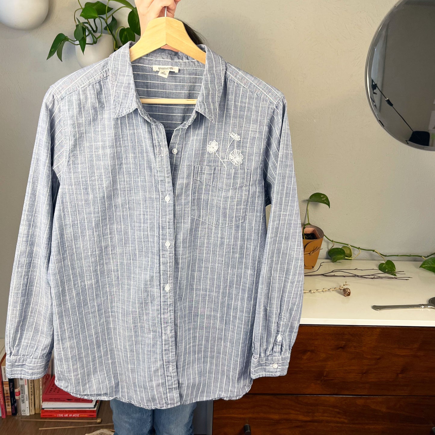 front view of a striped chambray shirt with white hand embroidered poppies coming out of the chest pocket being held up on a hanger in front of a table with books on it and a dresser and wall with plants on it