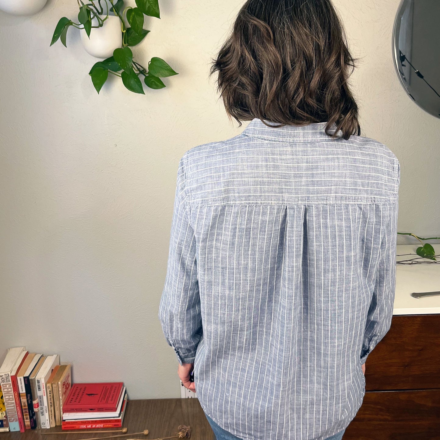 back view of a woman wearing a striped chambray button up shirt standing in front of a table with books on it and plants on a dresser and the wall