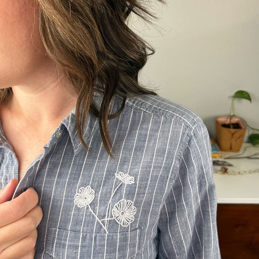 close up view of the front of a woman wearing a striped chambray button up shirt with three white hand embroidered poppies coming out of the chest pocket