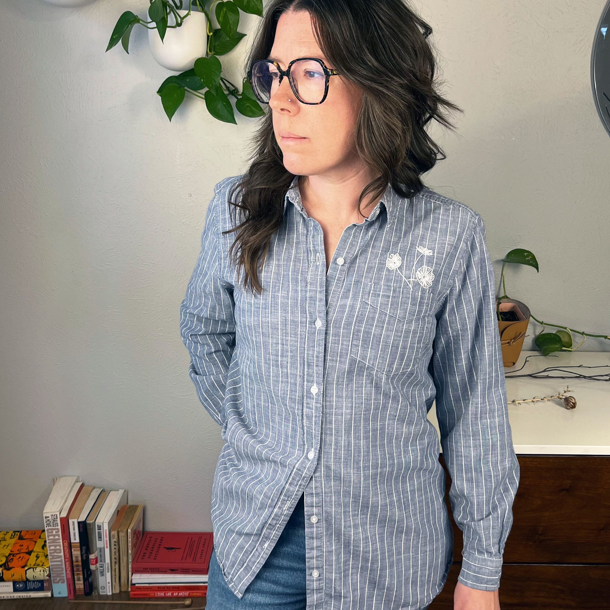 a woman wearing a striped chambray button up shirt with three white hand embroidered poppies coming out of the chest pocket standing in front of a table with books on it and plants on a dresser and the wall