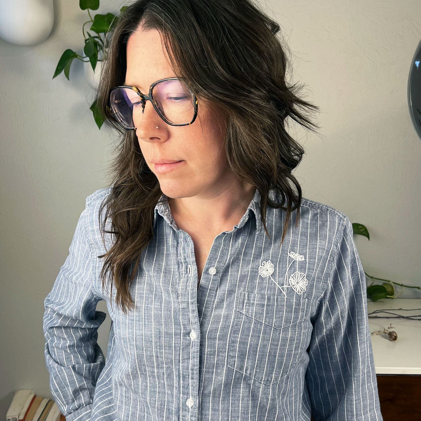 waist up view of a woman wearing a striped chambray button up shirt with three white hand embroidered poppies coming out of the chest pocket