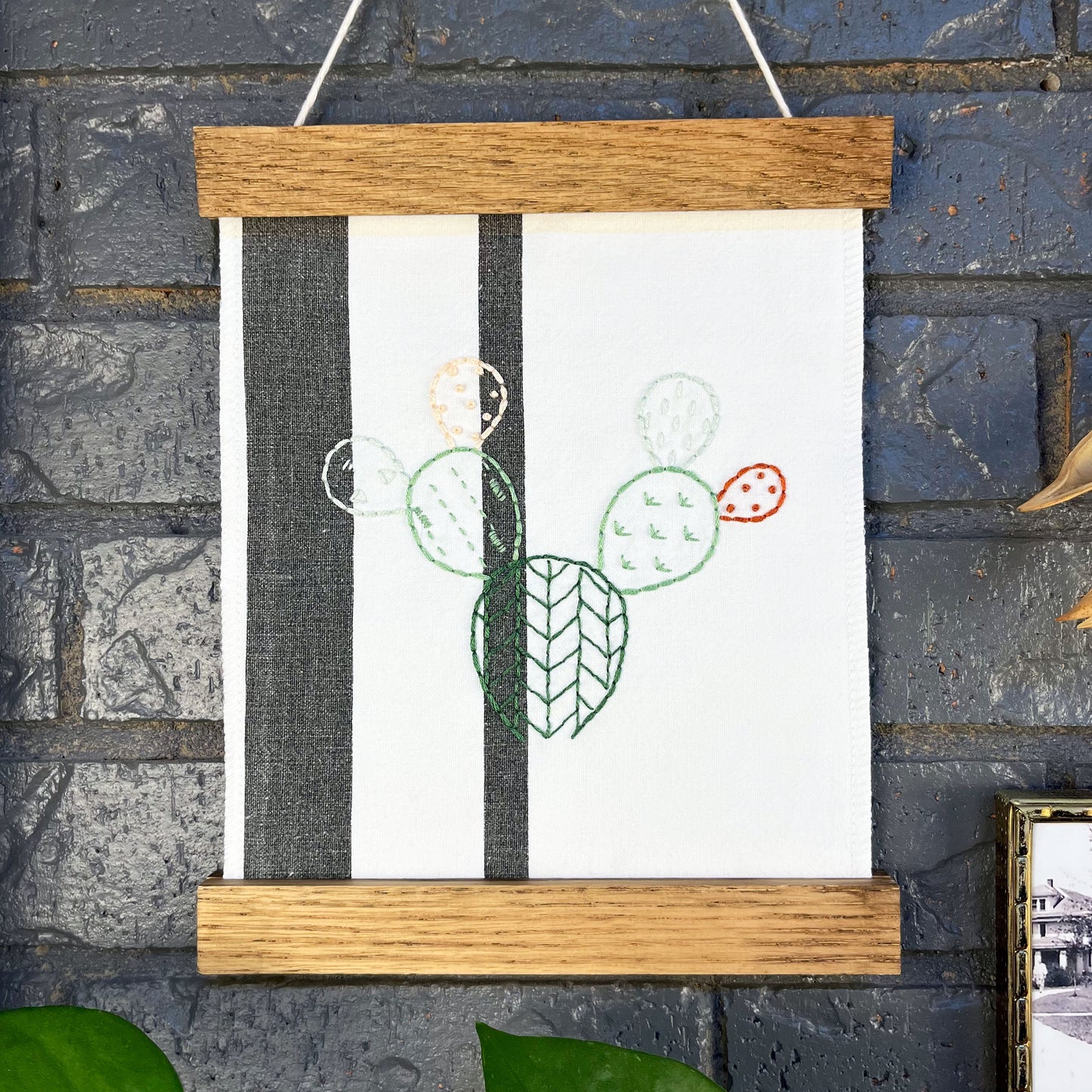 a small white fabric wall hanging with two black stripes in a magnetic wood frame, hand embroidered with the outline of a prickly pear cactus in greens peach and rust brown, each pad filled with different types of stitches, hanging on a brick wall near dried seed pods and a black and white photo in an antique brass frame