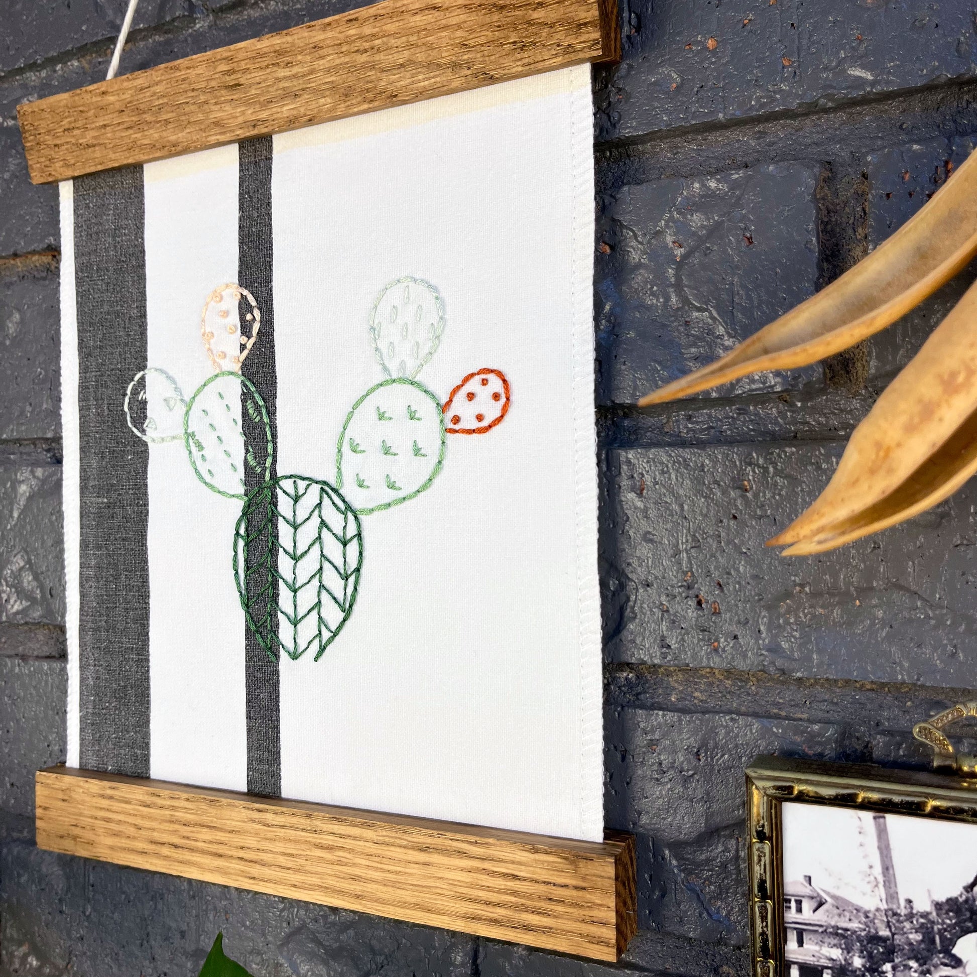 close up angled view of a small white fabric wall hanging with two black stripes in a magnetic wood frame, hand embroidered with the outline of a prickly pear cactus in greens peach and rust brown, each pad filled with different types of stitches, hanging on a brick wall near dried seed pods and a black and white photo in an antique brass frame