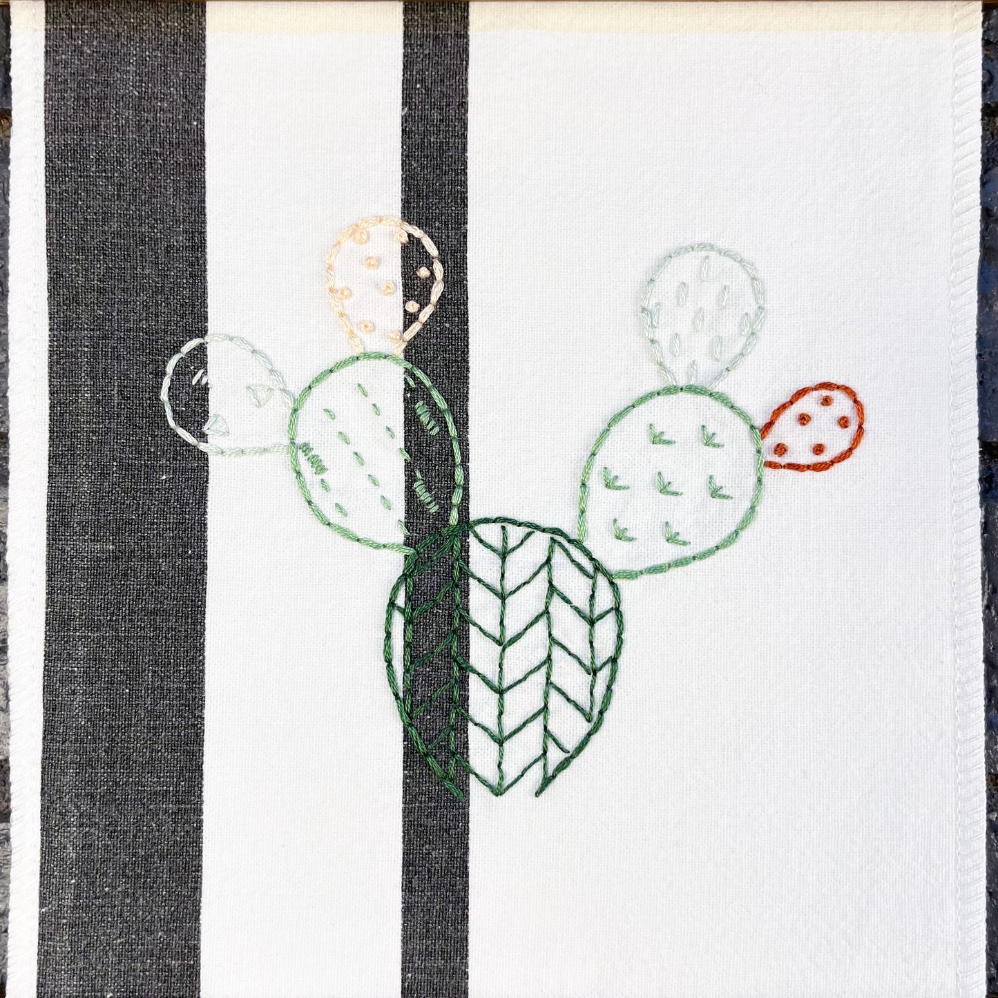 close up view of an outline of a prickly pear cactus hand embroidered in greens peach and rust brown on a white fabric with two black stripes, each pad filled with different types of stitches