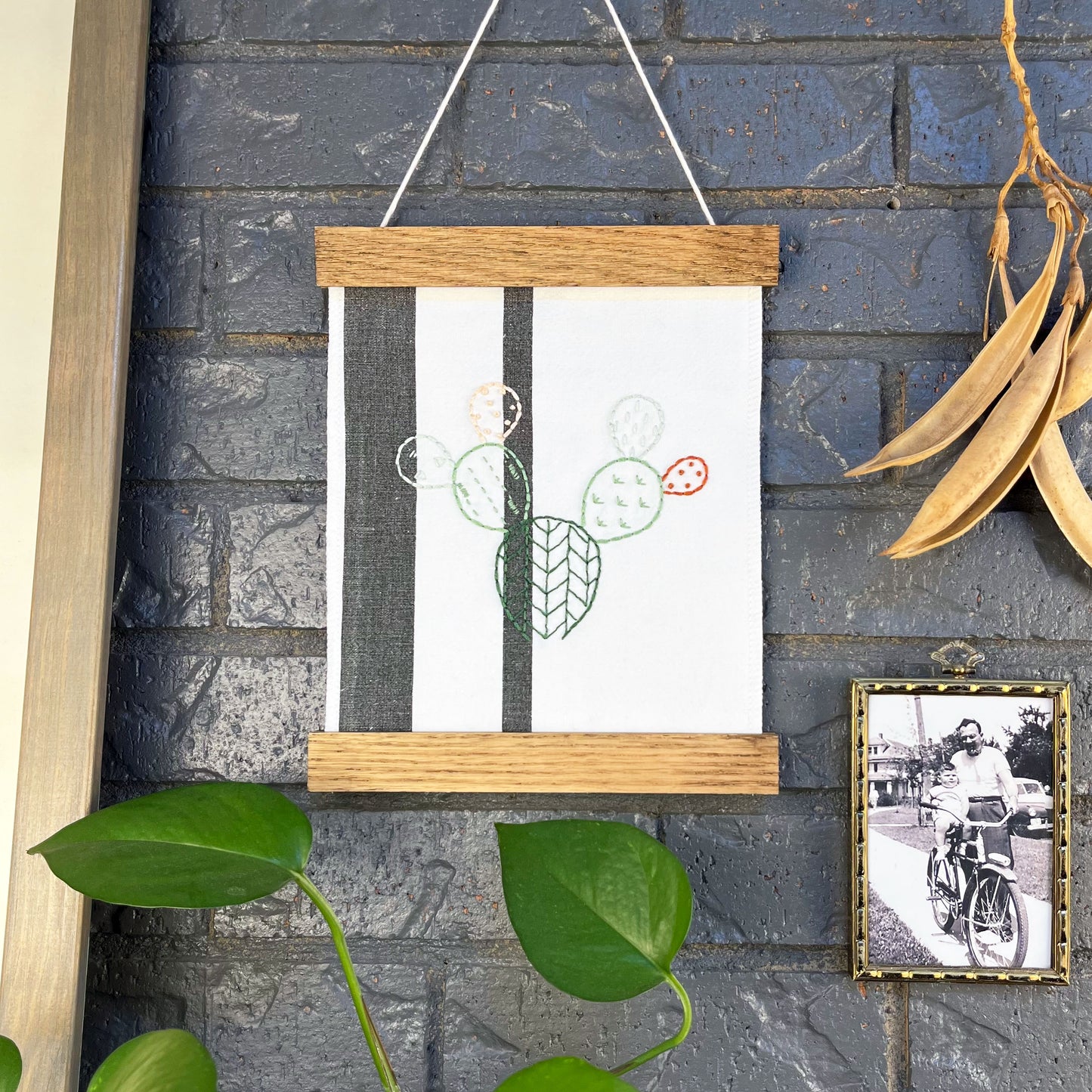 a small white fabric wall hanging with two black stripes in a magnetic wood frame, hand embroidered with the outline of a prickly pear cactus in greens peach and rust brown, each pad filled with different types of stitches, hanging on a brick wall near dried seed pods and a black and white photo in an antique brass frame