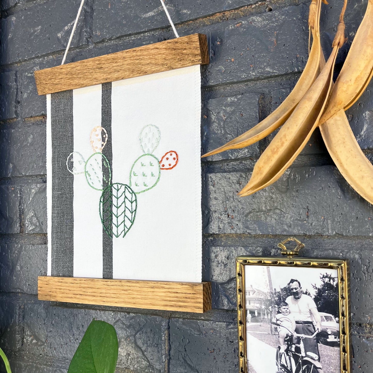 close up angled view of a small white fabric wall hanging with two black stripes in a magnetic wood frame, hand embroidered with the outline of a prickly pear cactus in greens peach and rust brown, each pad filled with different types of stitches, hanging on a brick wall near dried seed pods and a black and white photo in an antique brass frame