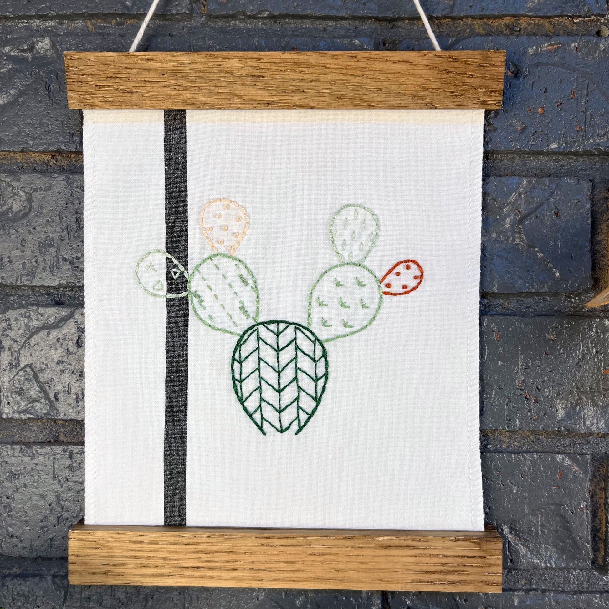 a small white fabric wall hanging with a black stripe in a magnetic wood frame, hand embroidered with the outline of a prickly pear cactus in greens peach and rust brown, each pad filled with different types of stitches, hanging on a grey brick wall 