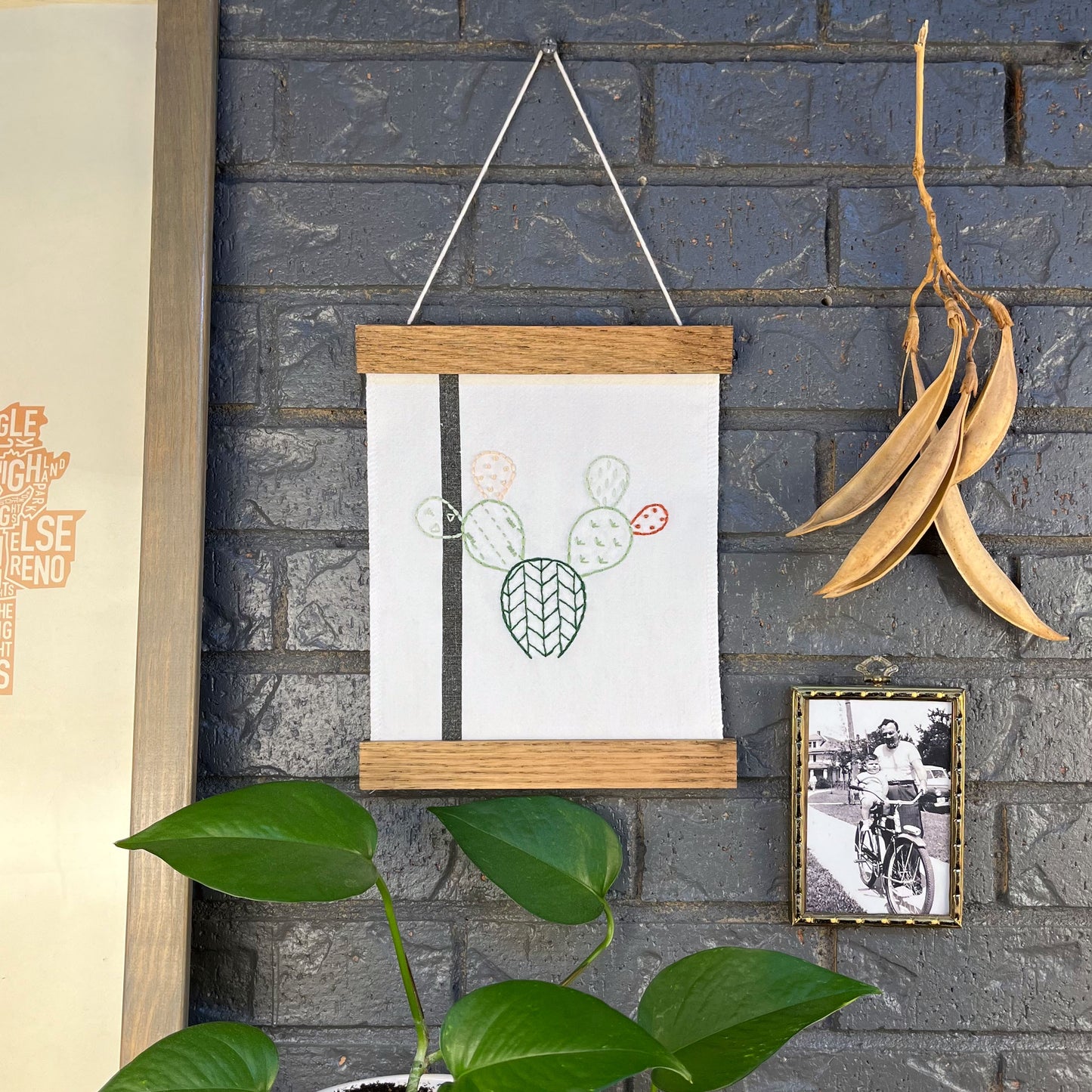 a small white fabric wall hanging with a black stripe in a magnetic wood frame, hand embroidered with the outline of a prickly pear cactus in greens peach and rust brown, each pad filled with different types of stitches, hanging on a brick wall near dried seed pods and an old black and white photo in an antique brass frame