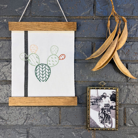 a small white fabric wall hanging with a black stripe in a magnetic wood frame, hand embroidered with the outline of a prickly pear cactus in greens peach and rust brown, each pad filled with different types of stitches, hanging on a brick wall near dried seed pods and an old black and white photo in an antique brass frame