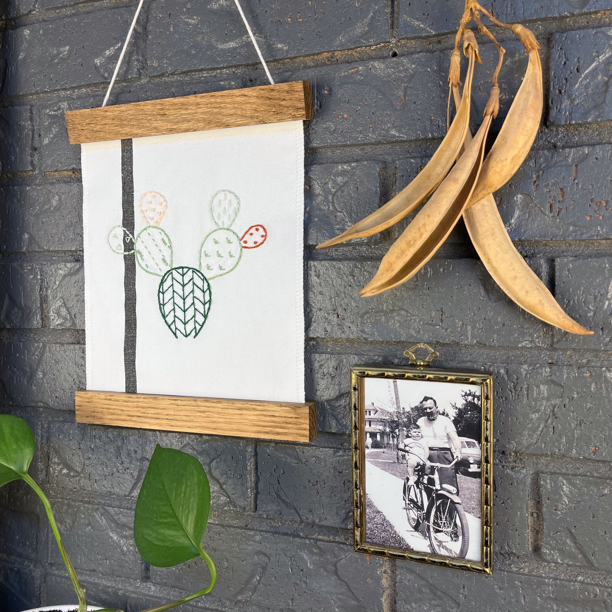 a small white fabric wall hanging with a black stripe in a magnetic wood frame, hand embroidered with the outline of a prickly pear cactus in greens peach and rust brown, each pad filled with different types of stitches, hanging on a brick wall near dried seed pods and an old black and white photo in an antique brass frame