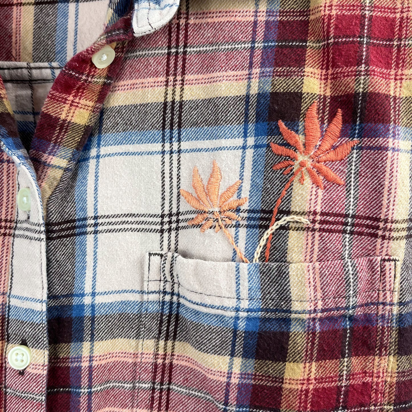 close up view of a peach and maroon plaid flannel with two hand embroidered flowers in peach and coral coming out of the chest pocket