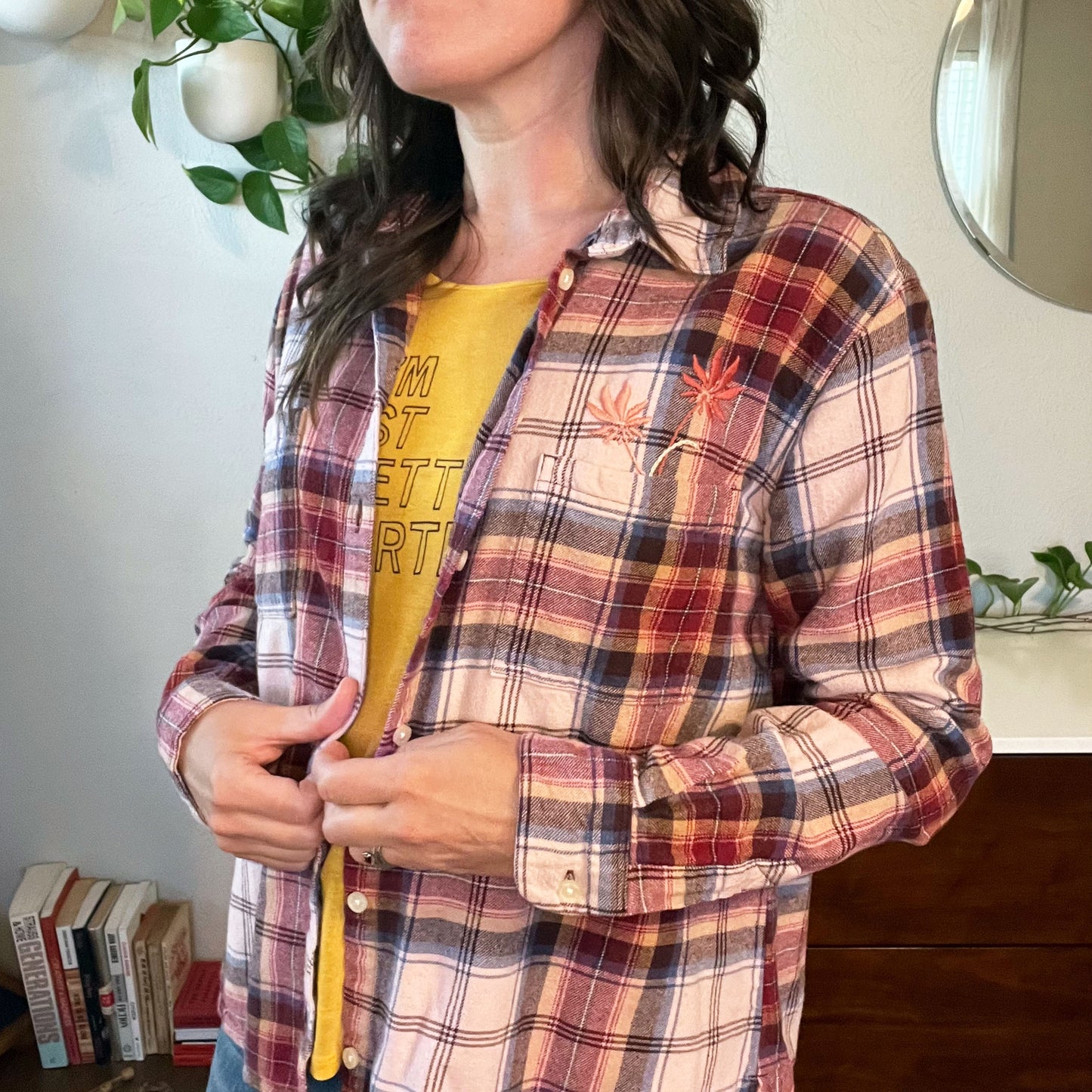 a woman wearing a peach and maroon plaid flannel with two hand embroidered flowers in peach and coral coming out of the chest pocket with books plants a white and wood dresser and a round mirror in the background