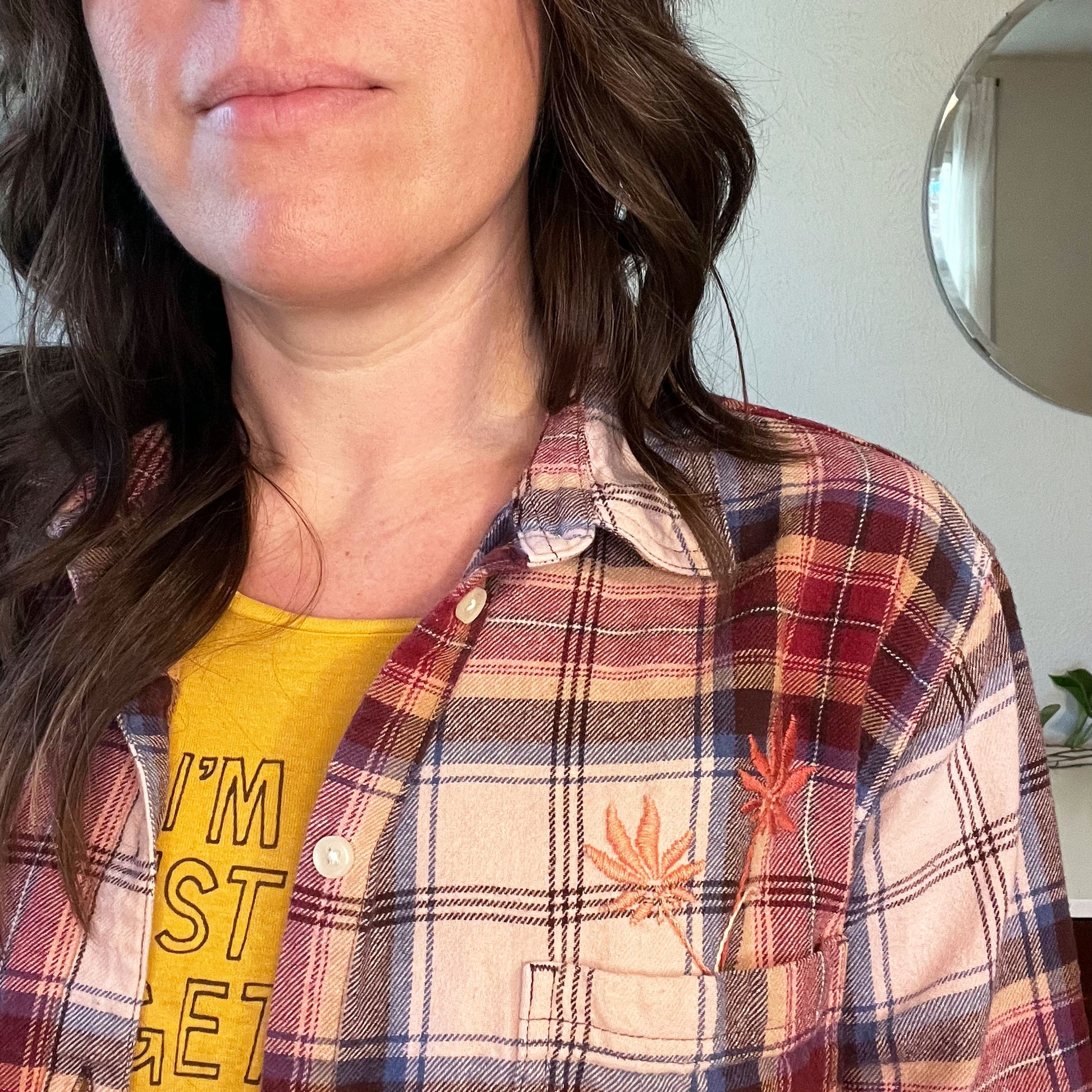 close up view of a woman wearing a peach and maroon plaid flannel with two hand embroidered flowers in peach and coral coming out of the chest pocket with a round mirror in the background