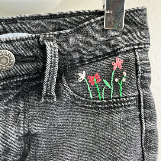close up view of black jean shorts front pocket with hand embroidered flowers in greens and pinks, using chainstitch, stem stitch, and french knots