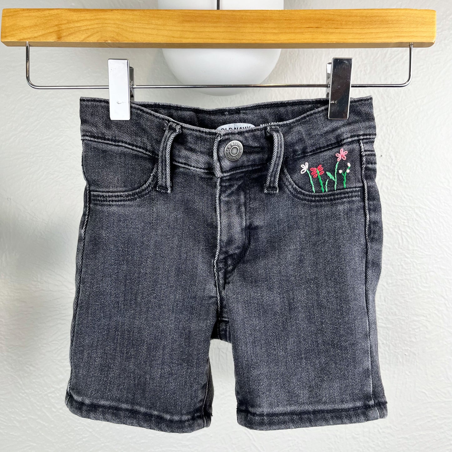 black jean shorts hanging from a wood hanger, one of the front pockets has hand embroidered flowers in greens and pinks, using chainstitch, stem stitch, and french knots