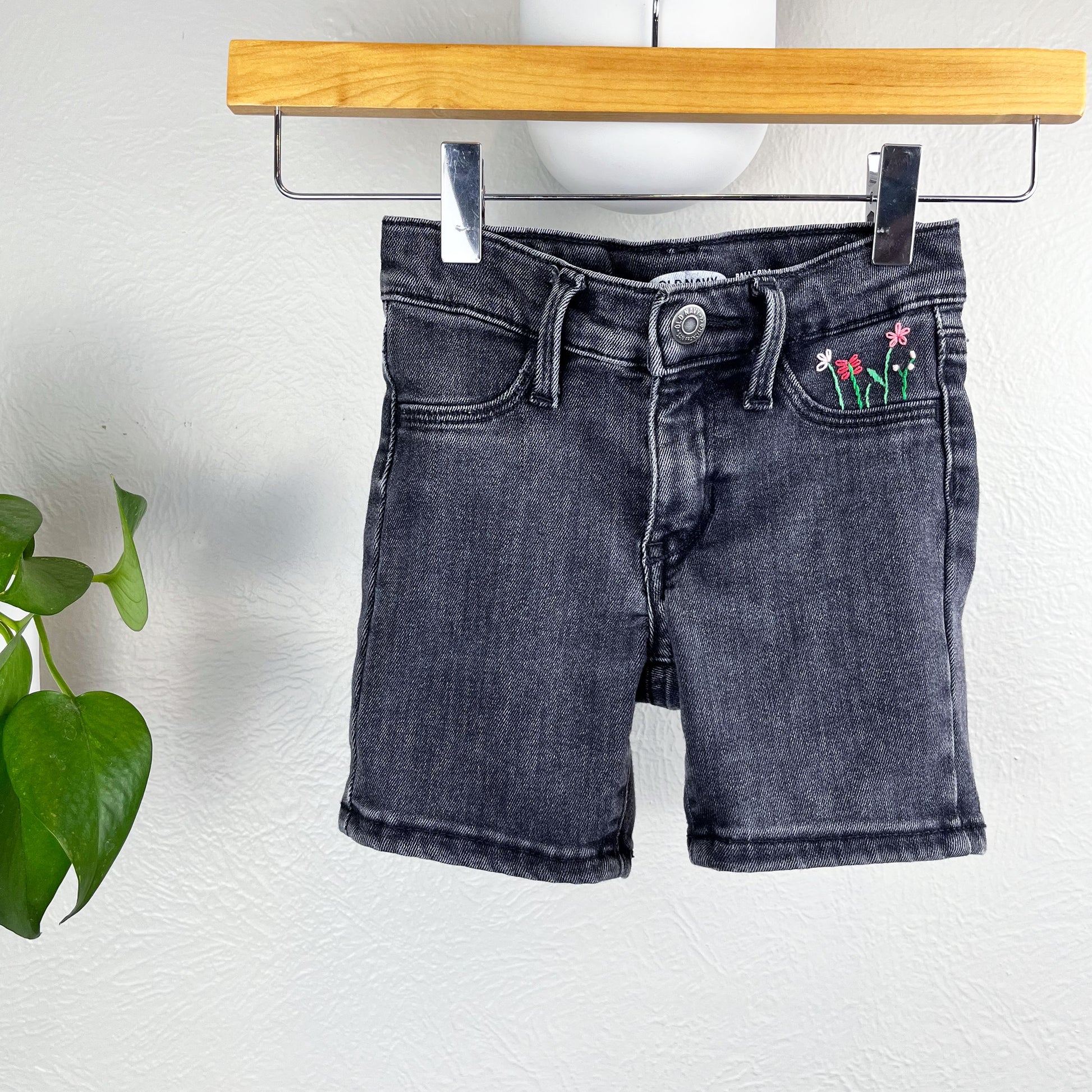 black jean shorts hanging from a wood hanger next to a pothos plant, one of the front pockets has hand embroidered flowers in greens and pinks, using chainstitch, stem stitch, and french knots