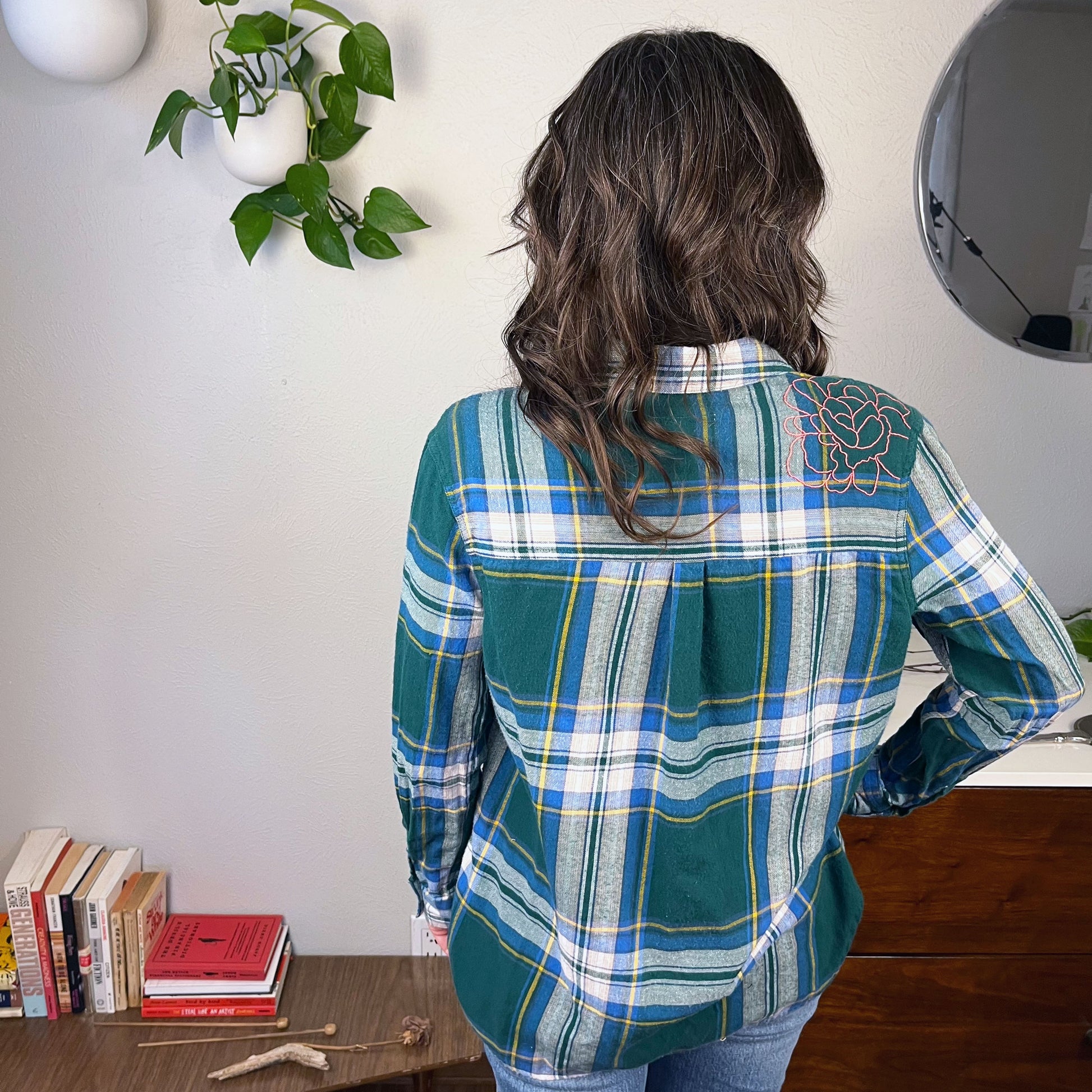 back view of a woman wearing a green and blue plaid flannel with an outline of a large peony hand embroidered in pink on the shoulder with books and plants in the background