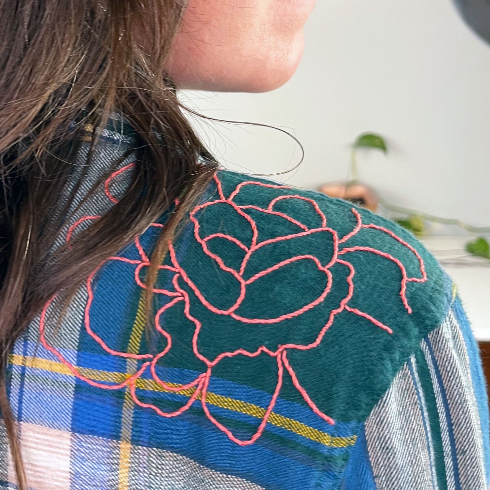 close up view of the back shoulder of a woman wearing a green and blue plaid flannel with an outline of a large peony hand embroidered in pink on the shoulder