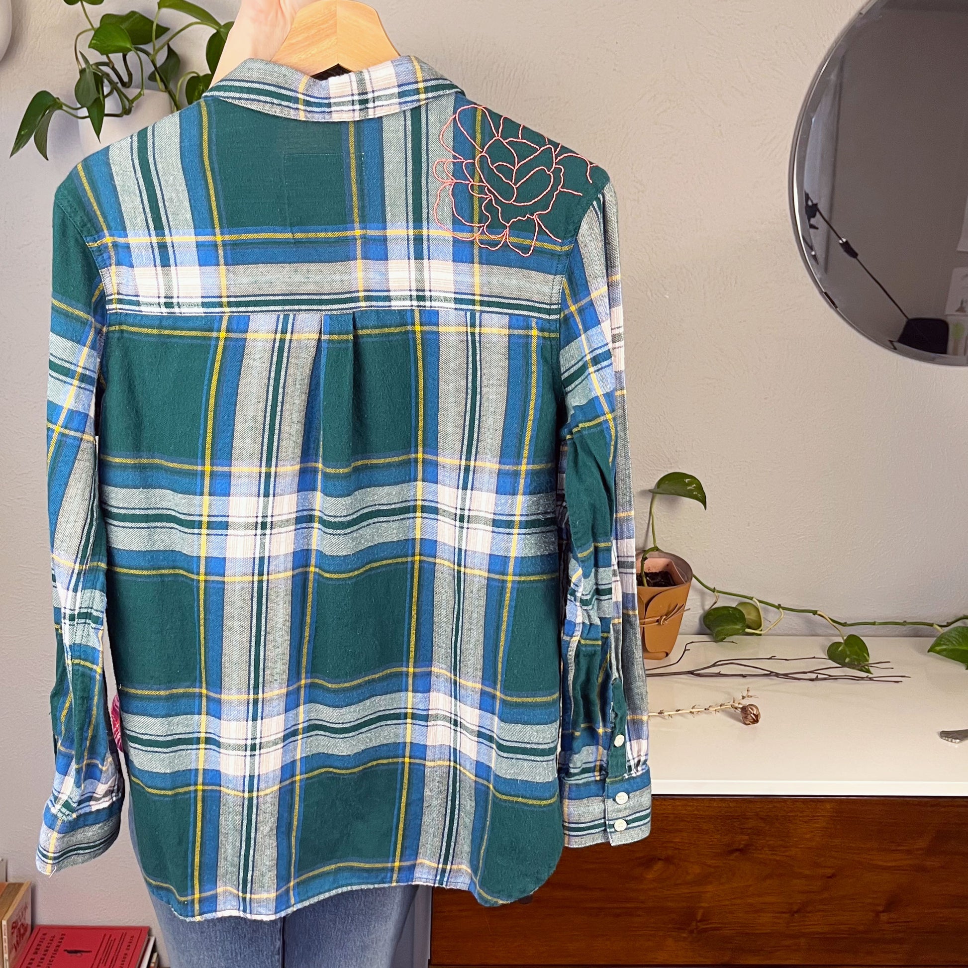 back view of a green and blue plaid flannel with an outline of a large peony hand embroidered in pink on the shoulder hanging on a hanger with books and plants in the background