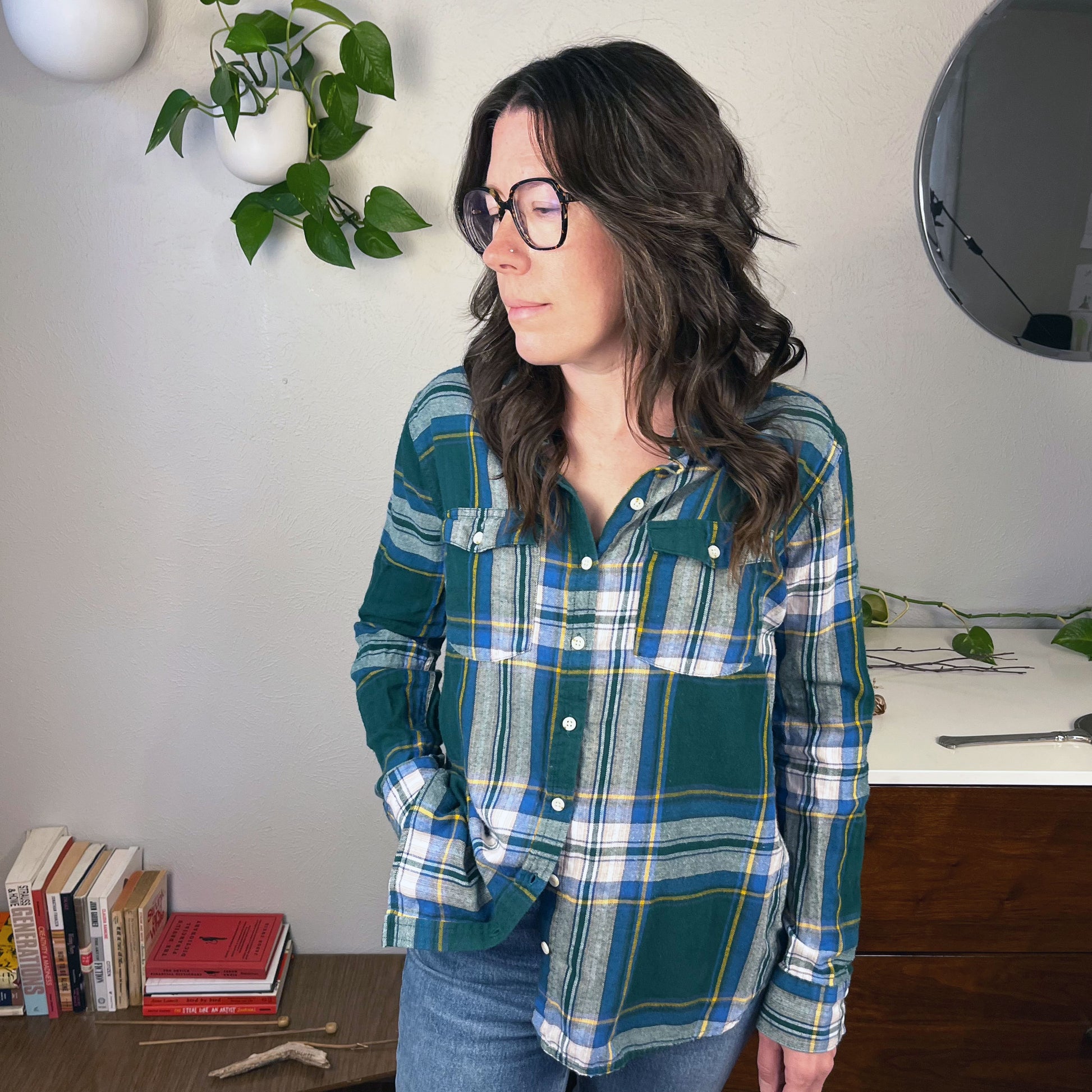 a white woman with glasses wearing a green and blue plaid flannel with books and plants in the background