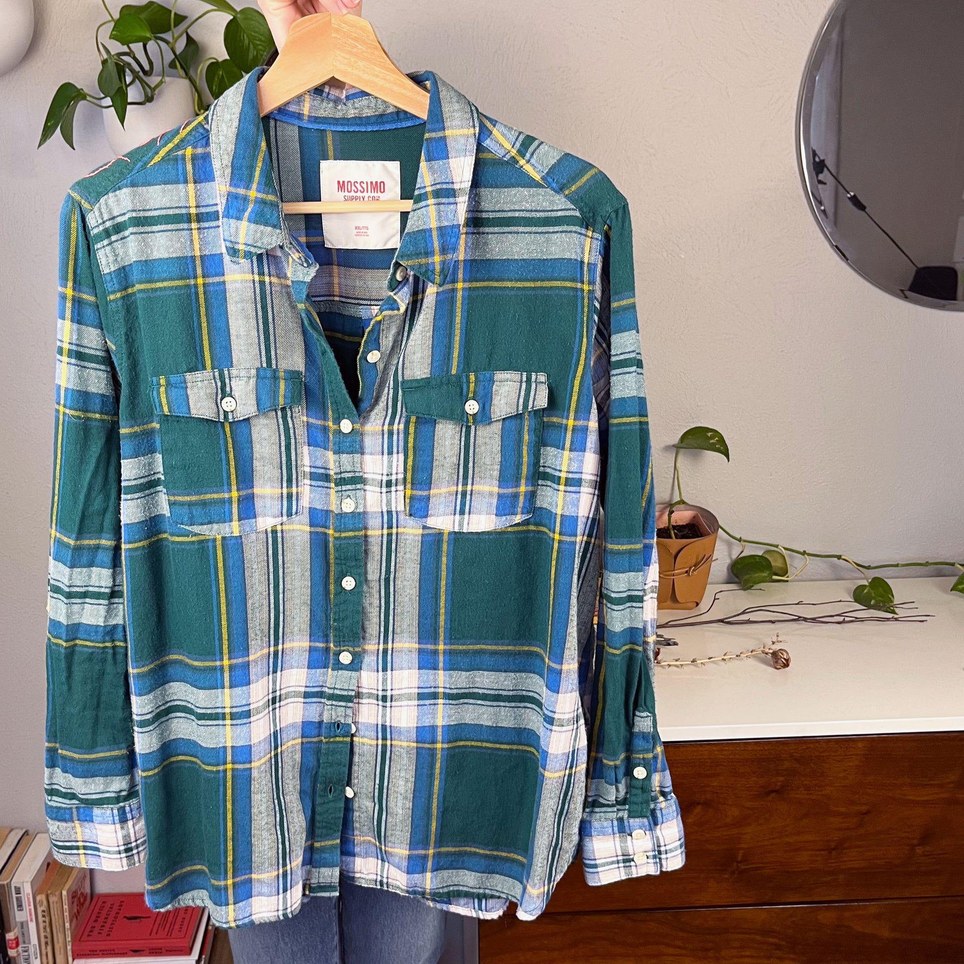 front view of a green and blue plaid flannel with an outline of a large peony hand embroidered in pink on the shoulder hanging on a hanger with books and plants in the background