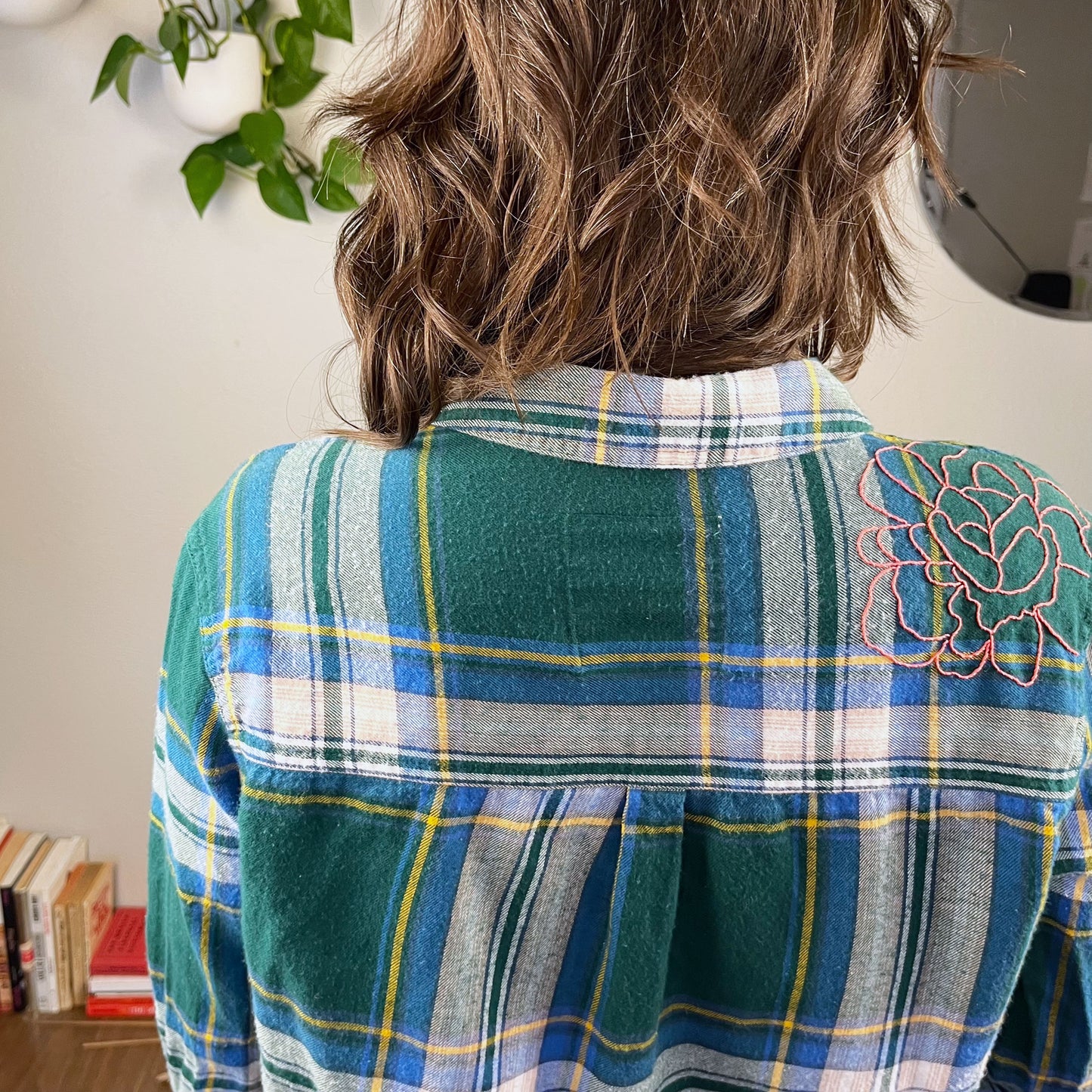 back view of a woman wearing a green and blue plaid flannel with an outline of a large peony hand embroidered in pink on the shoulder with books and plants in the background