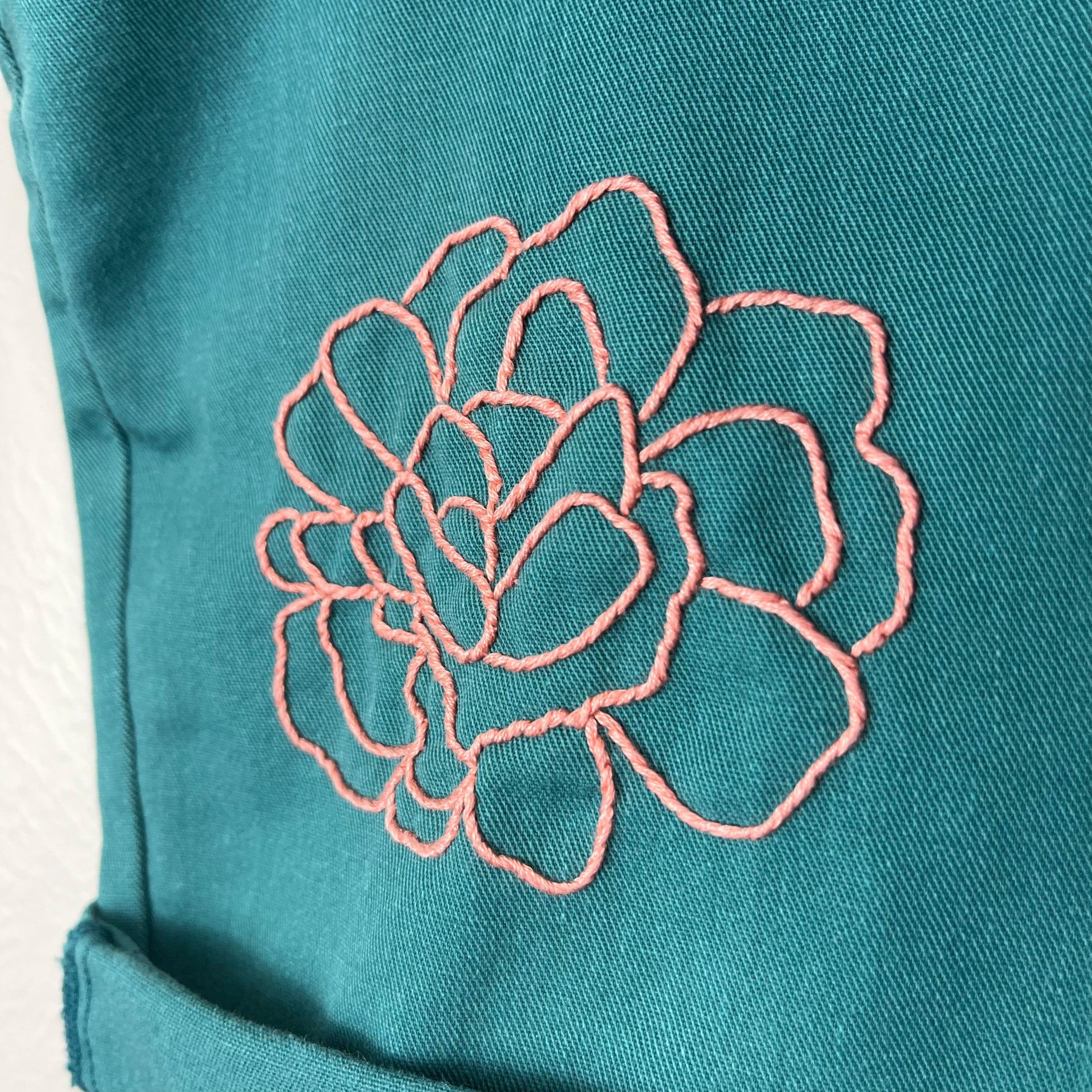 close up view of an outline of a peony hand embroidered in coral peach floss on green shorts