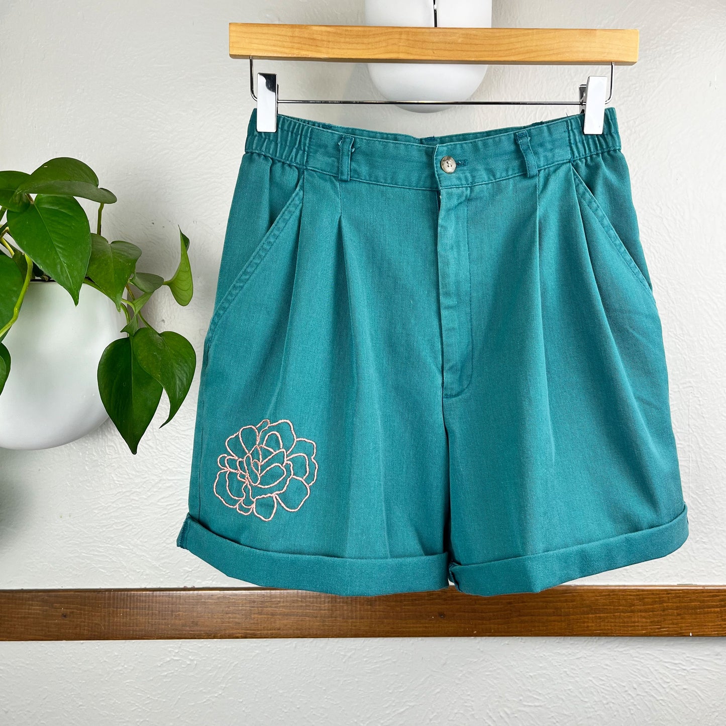 a pair of pleated green high waisted shorts, with an outline of a peony hand embroidered in coral peach floss near the front right leg opening, hanging near a pothos plant