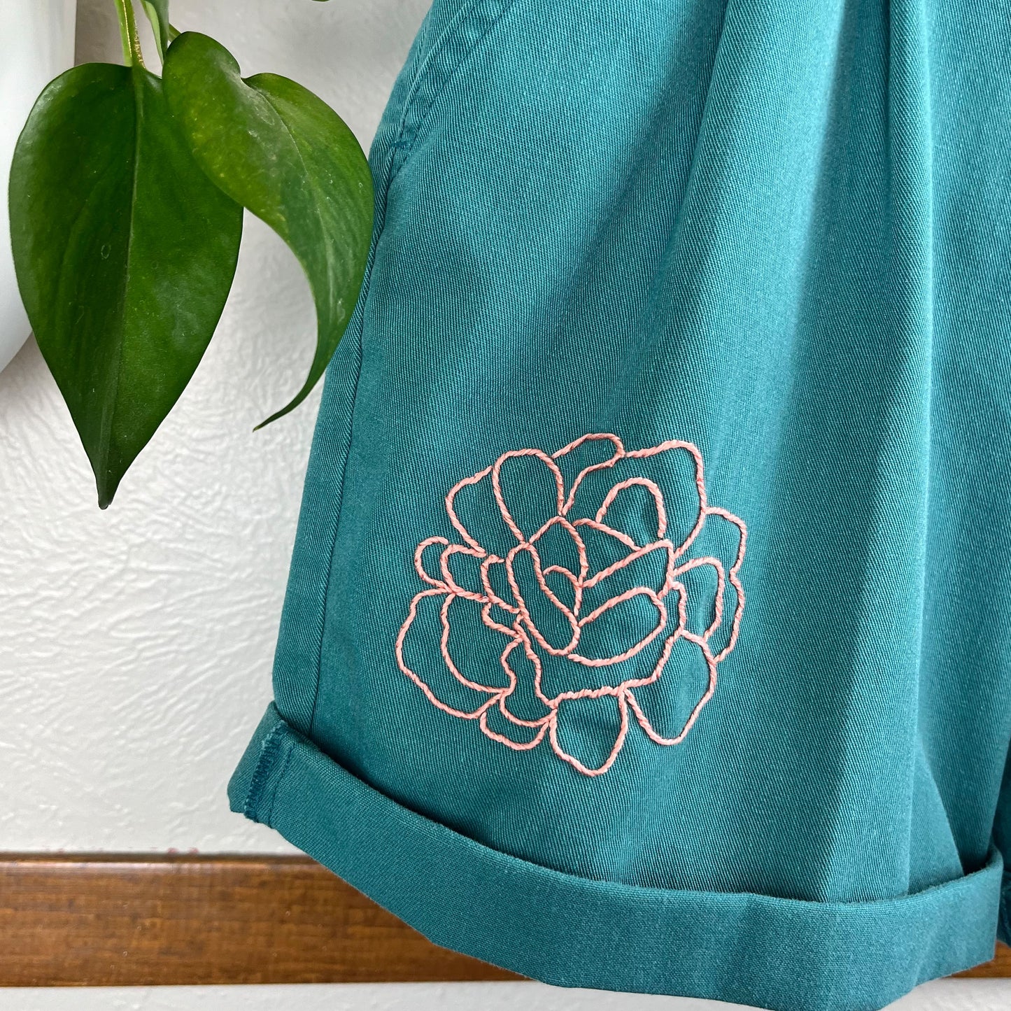 close up view of the bottom right leg of a pair of pleated green shorts, with an outline of a peony hand embroidered in coral peach floss, hanging near a pothos plant