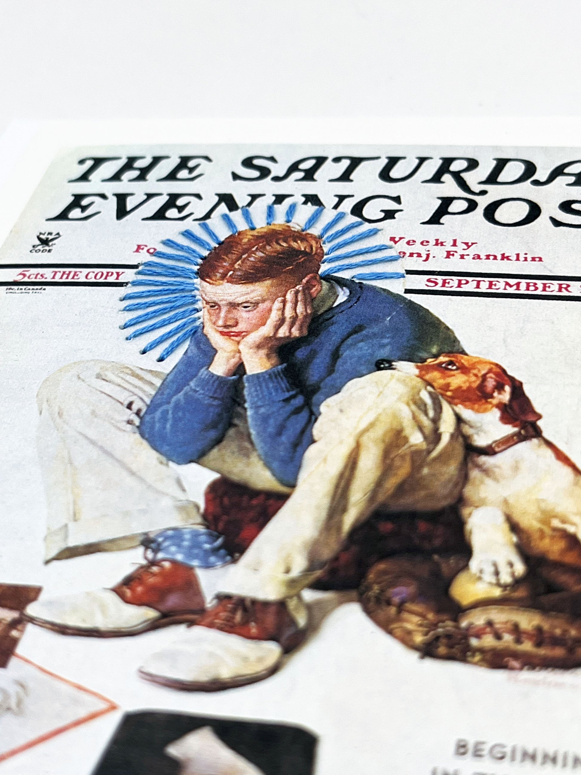 greeting card with a print of an issue of The Saturday Evening Post, a picture of a boy looking distraught with his dog while looking at magazine covers, a blue hand embroidered circle of rays around his head