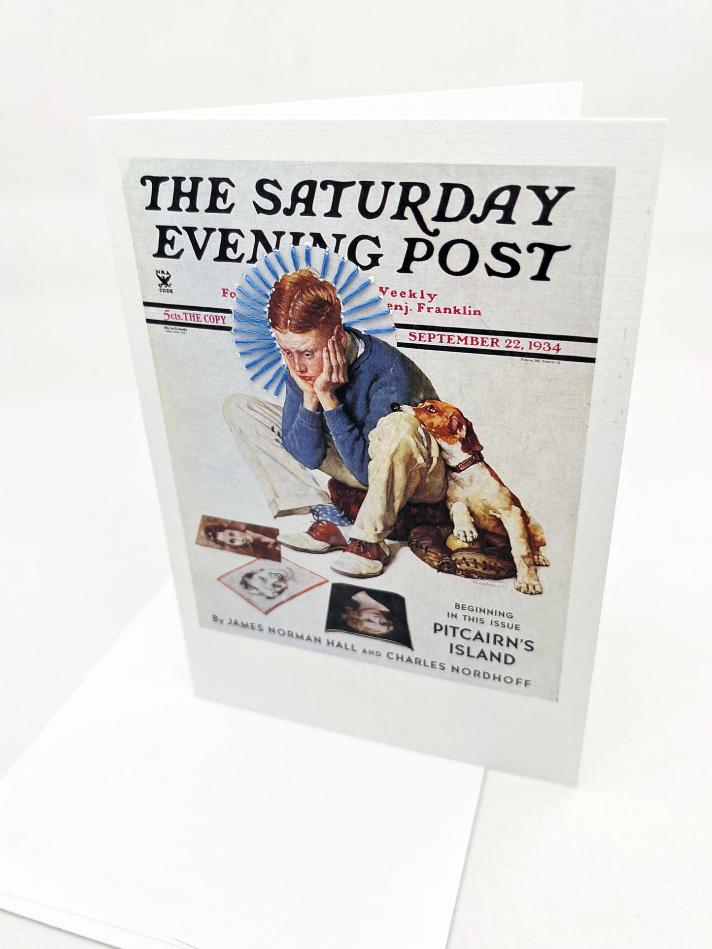 greeting card with a print of an issue of The Saturday Evening Post, a picture of a boy looking distraught with his dog while looking at magazine covers, a blue hand embroidered circle of rays around his head