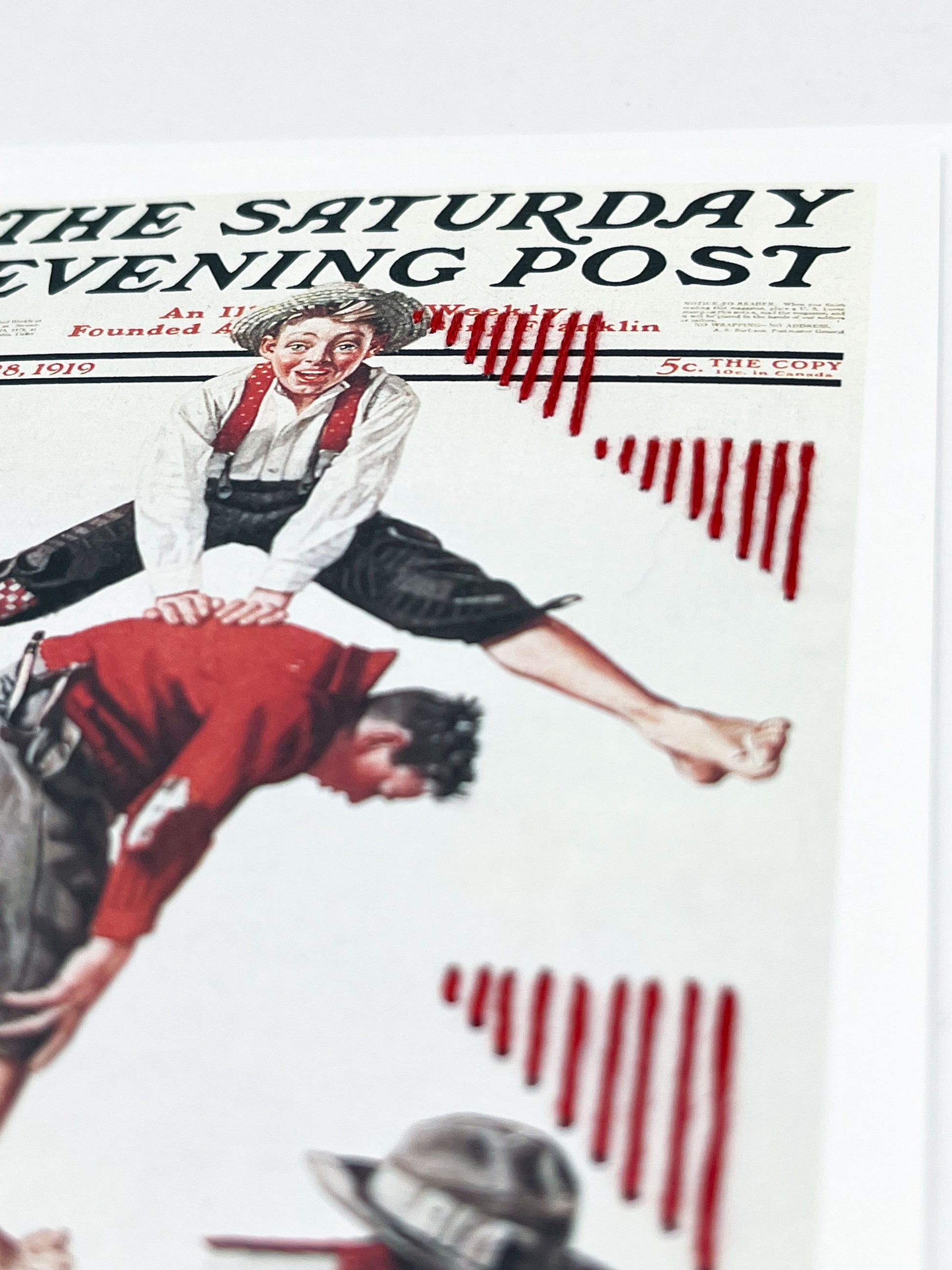 greeting card with a print of an issue of The Saturday Evening Post, a picture of a boy jumping over another boy, a with triangles made of red stripes hand embroidered around them