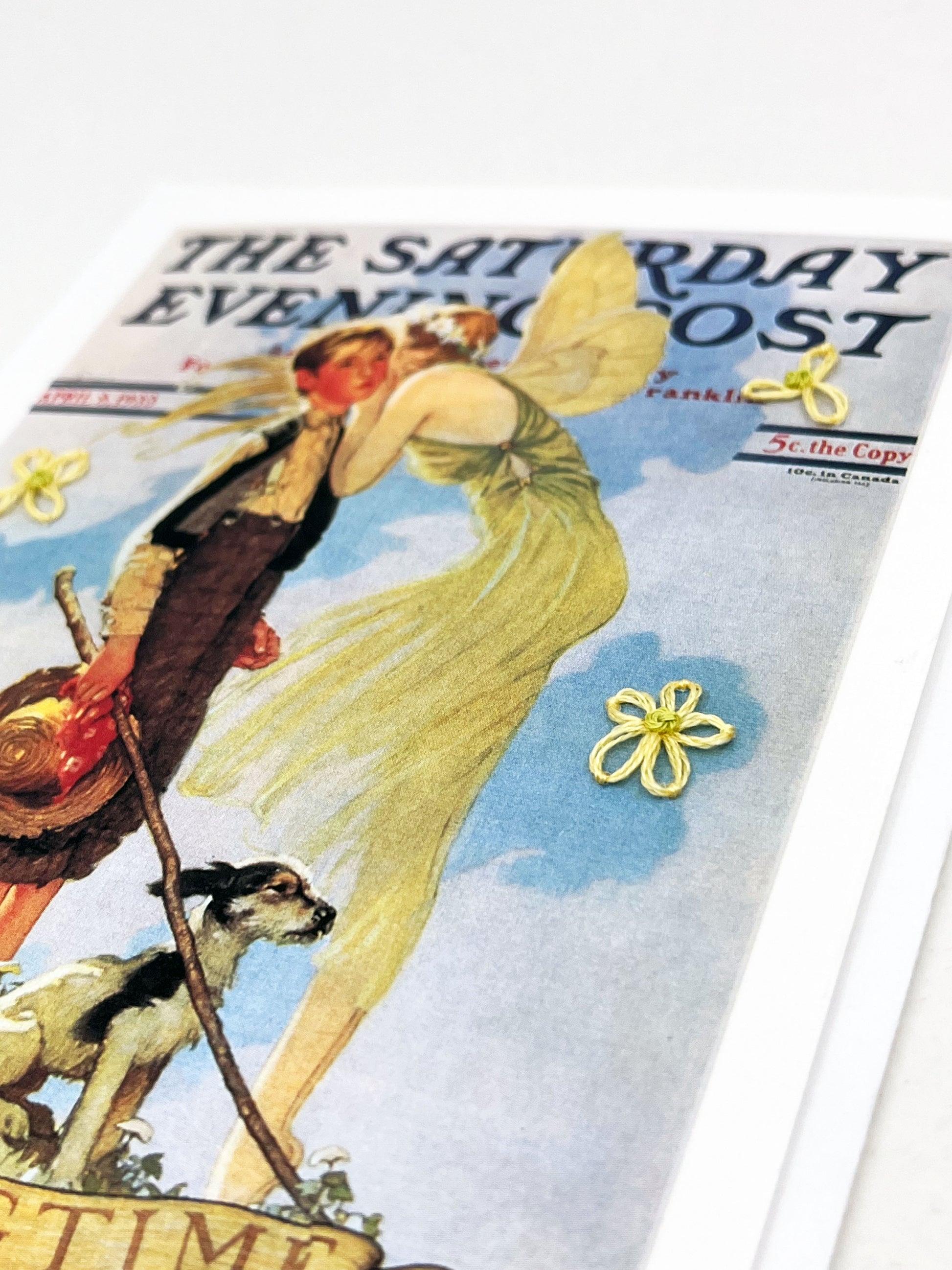 greeting card with a print of an issue of The Saturday Evening Post, a picture of a fairy whispering in a boys ear, a with hand embroidered flowers around them