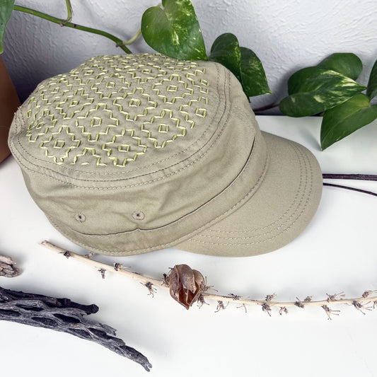 tan cadet hat with persimmon sashiko pattern stitched in chartreuse thread on the top, sitting on a dresser with a plant vine and dried flowers around it