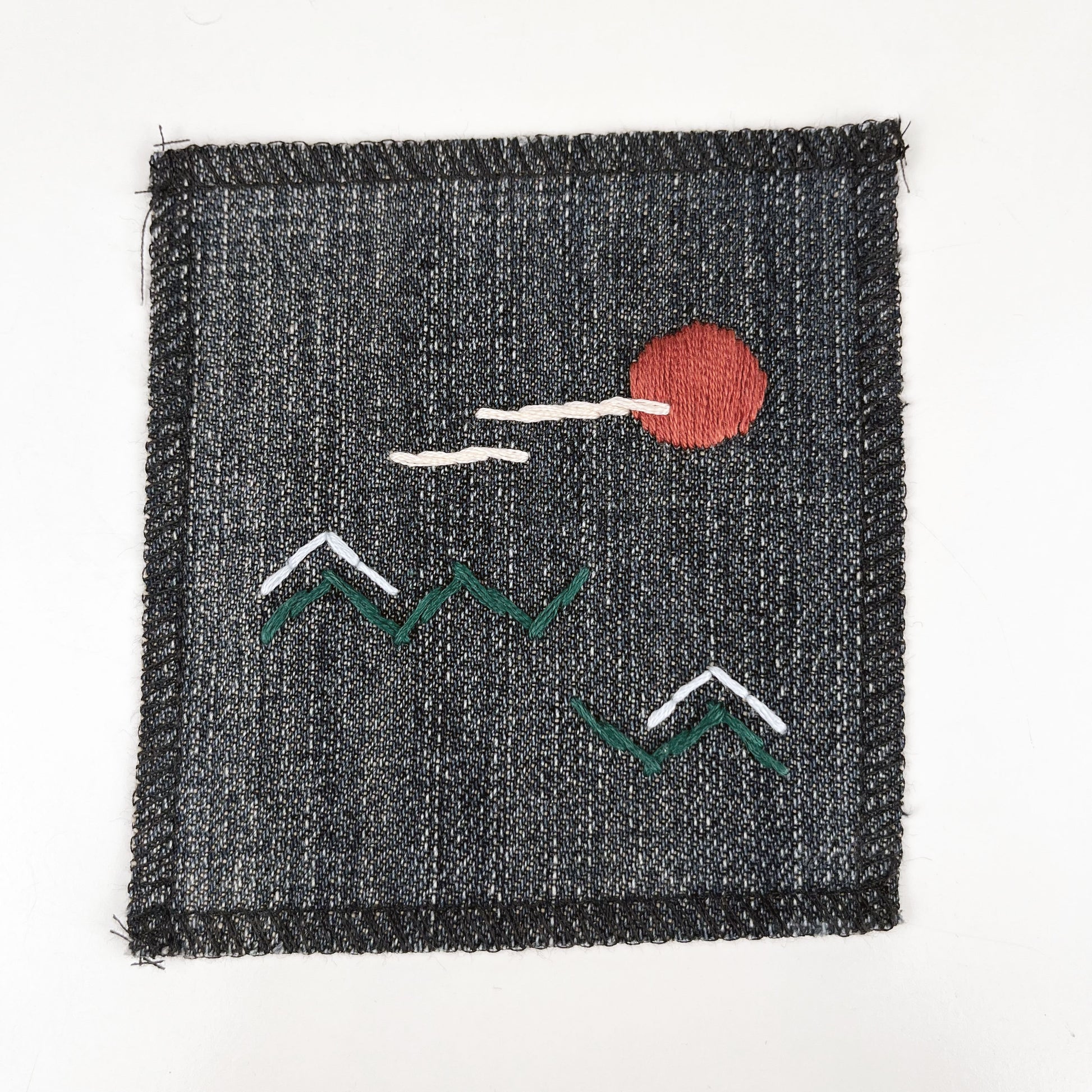 a square denim patch, with an abstract image hand stitched on them of a rust colored sun, with lines of peach clouds in front of it, over pine green zigzags as mountains, some with a line of light blue snow over them, overlocked edges