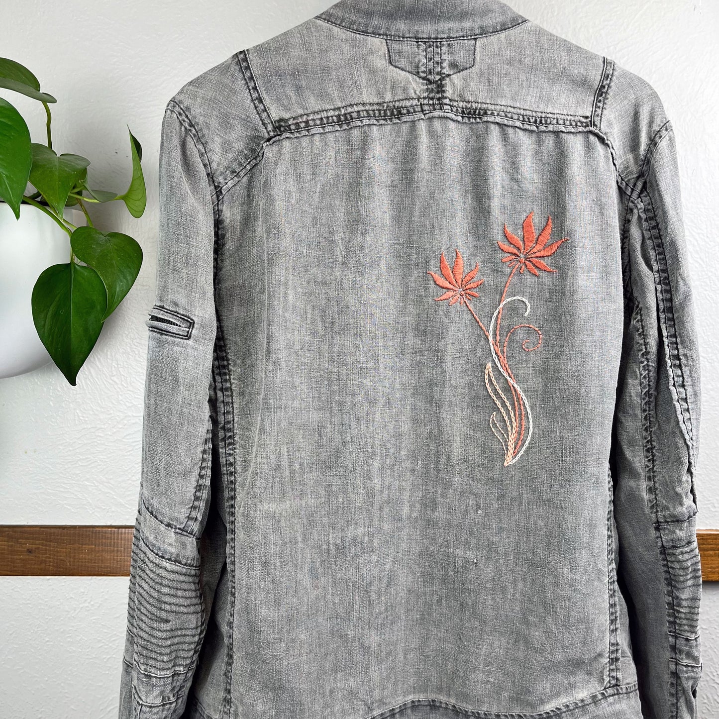 back of a grey linen moto jacket with long coral and peach  flowers hand embroidered on the right side, hanging on a wall next to a pothos plant