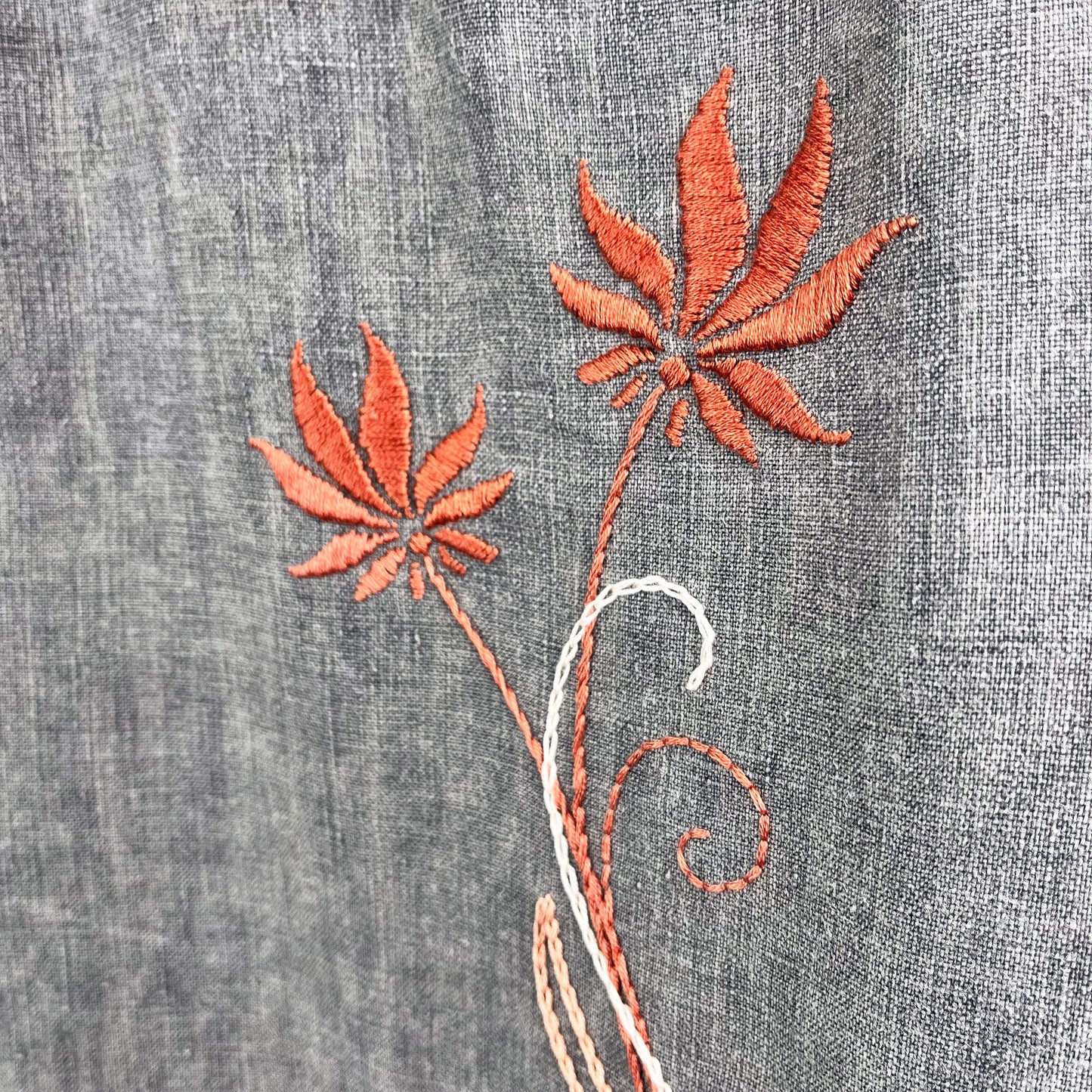 close up view of two hand embroidered coral flowers with leaves and flourishes in peach on grey linen, stitches used are backstitch, chainstitch, stem stitch, and satin fill