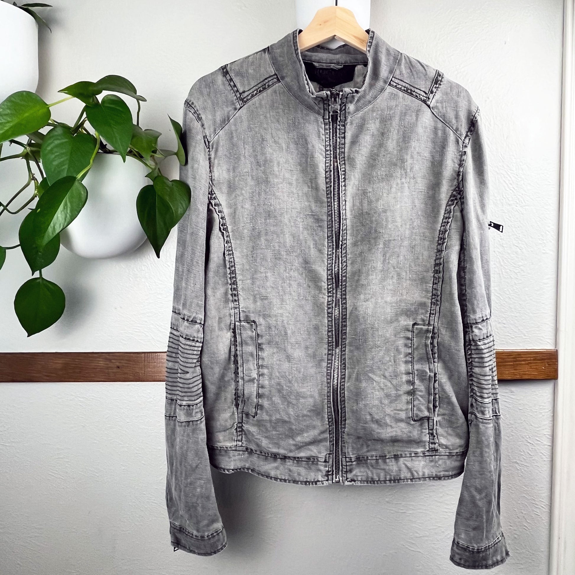 front side of a grey linen moto jacket, hanging on a wall next to a pothos plant