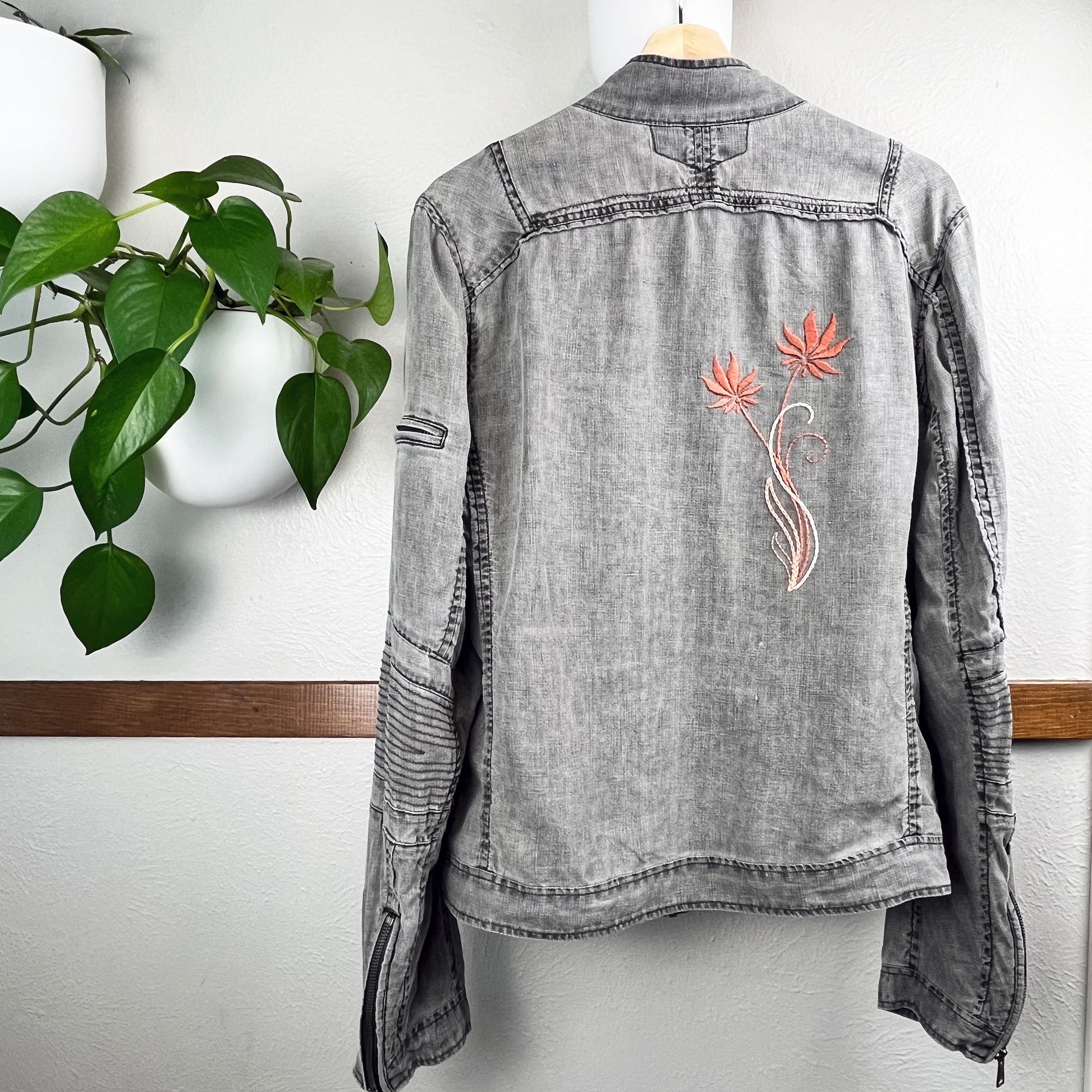 back of a grey linen moto jacket with long coral and peach  flowers hand embroidered on the right side, hanging on a wall next to a pothos plant