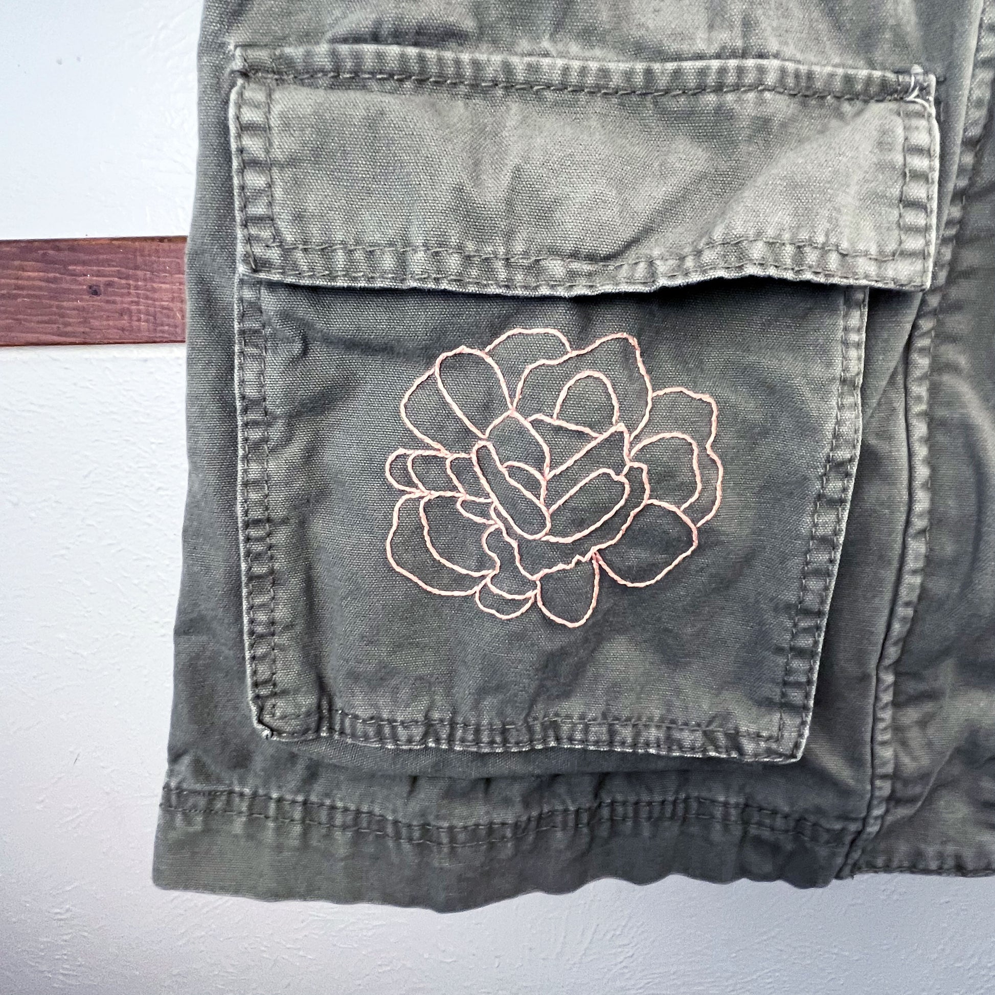 close up view of a right hip pocket of a military green jacket with an outline of a peony hand embroidered in peach on it