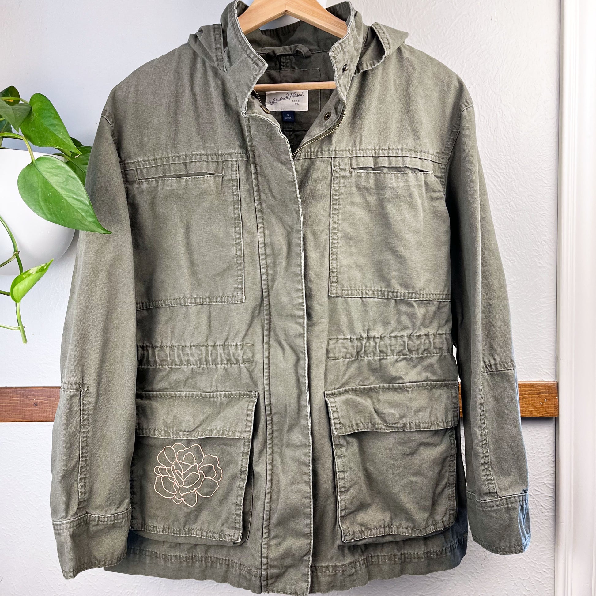 a military green cotton parka with many pockets and a hood, the right hip pocket has an outline of a peony hand embroidered in peach on it, hanging on a hanger on a wall next to a pothos plant